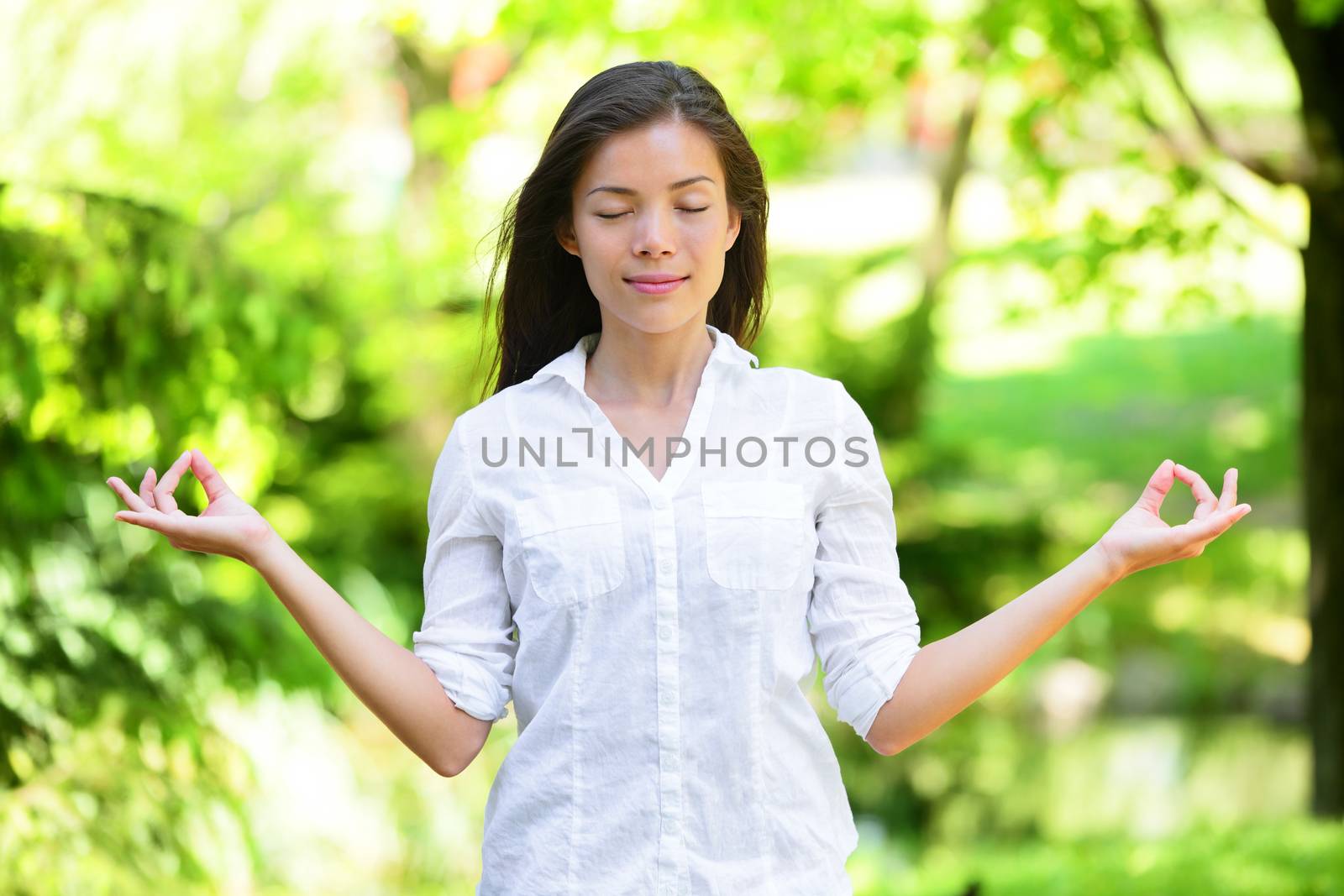 Beautiful young woman meditating in park. Attractive female in casuals is standing in lotus position. Peaceful female with eyes closed is practicing yoga in nature.