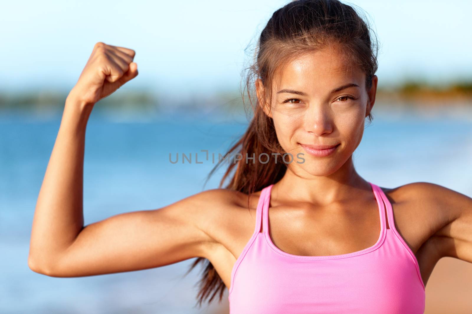 Confident sporty woman flexing muscles on beach. Beautiful young is wearing pink sports bra. Female is showing her strength and healthy lifestyle on sunny day.