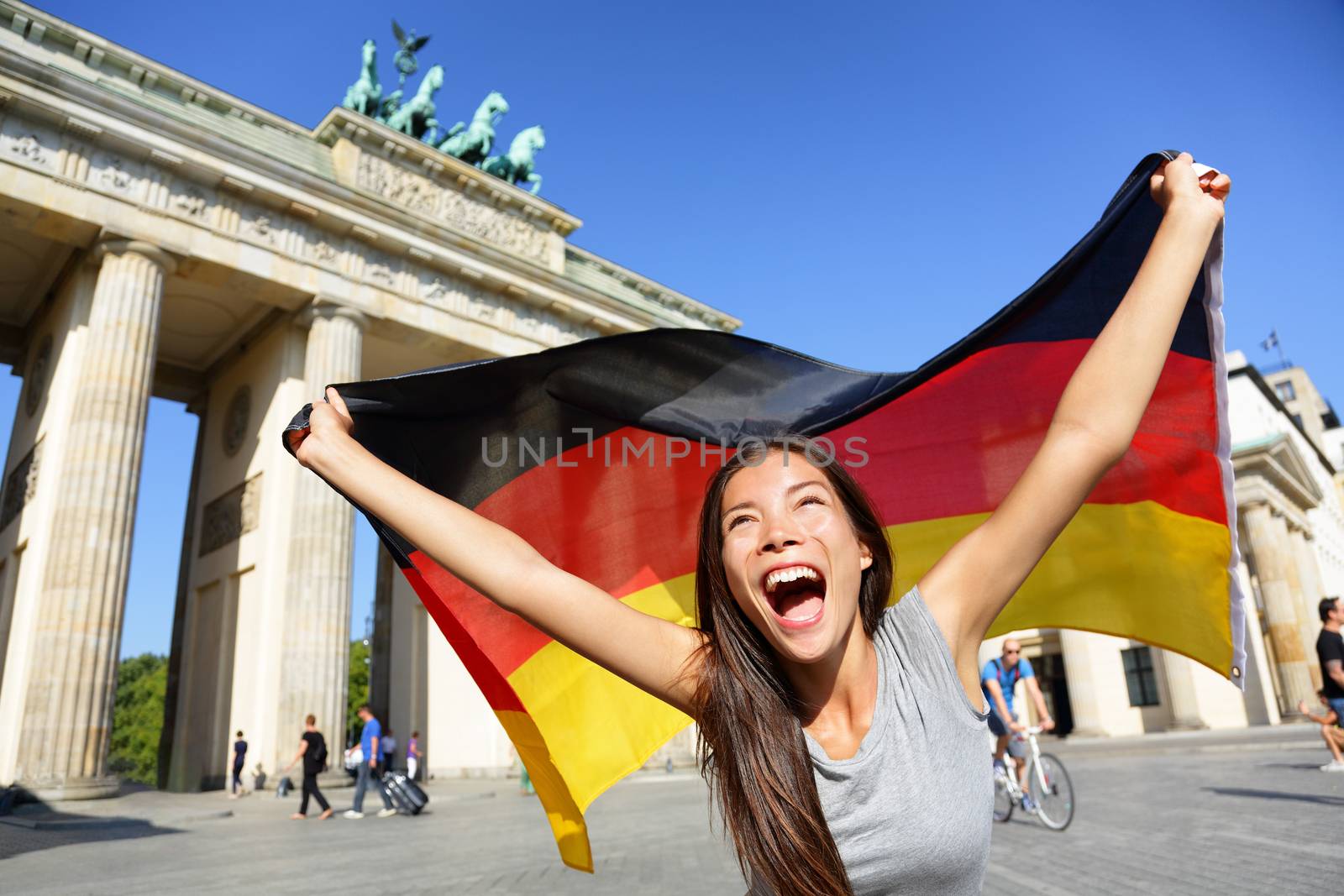 Excited young woman screaming while waving German flag in front of Brandenburg Gate. Cheerful female is enjoying her vacation in Berlin. Beautiful woman is celebrating her holidays.