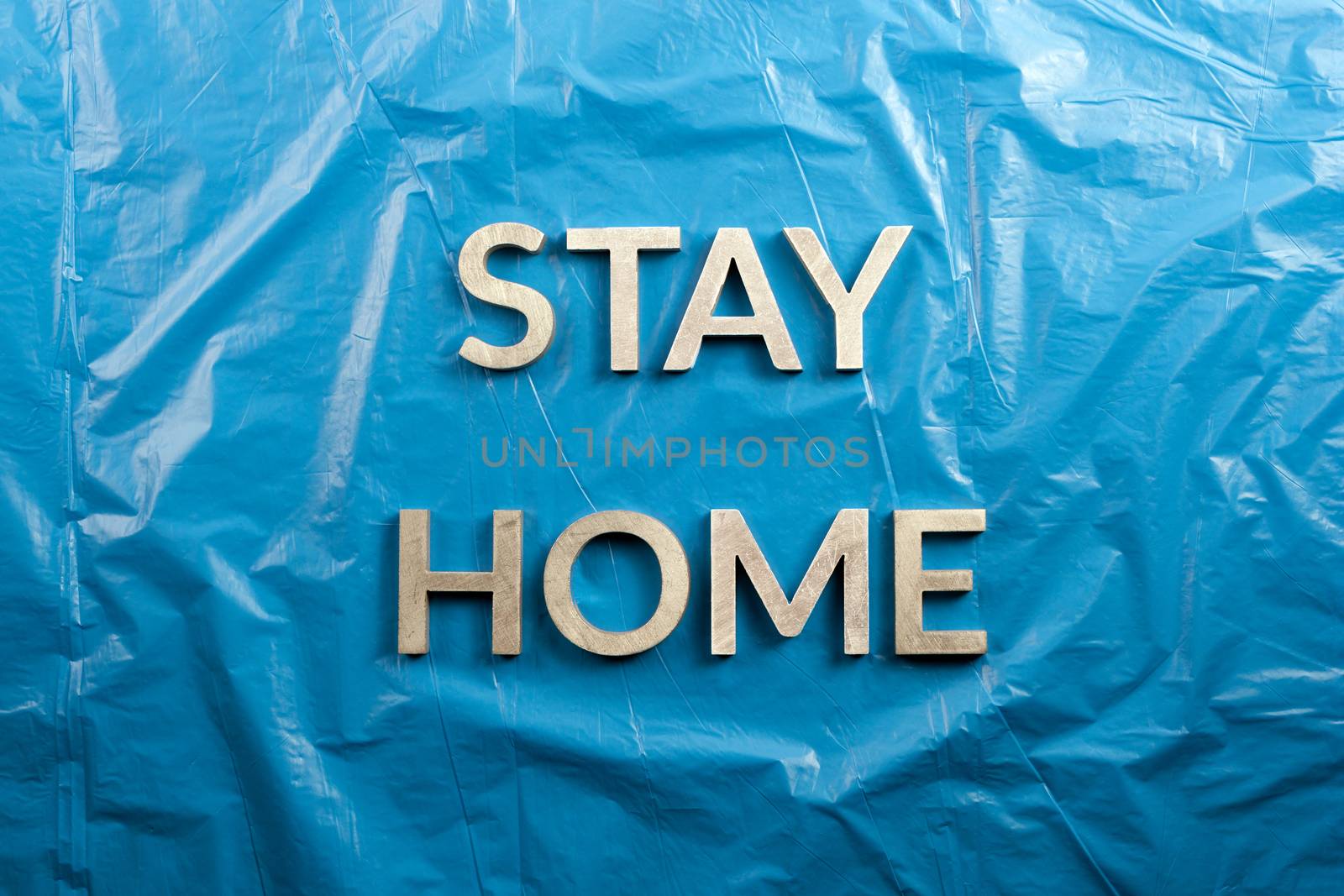 the words stay home laid with silver metal letters on crumpled blue plastic background in flat lay perspective, directly above composition