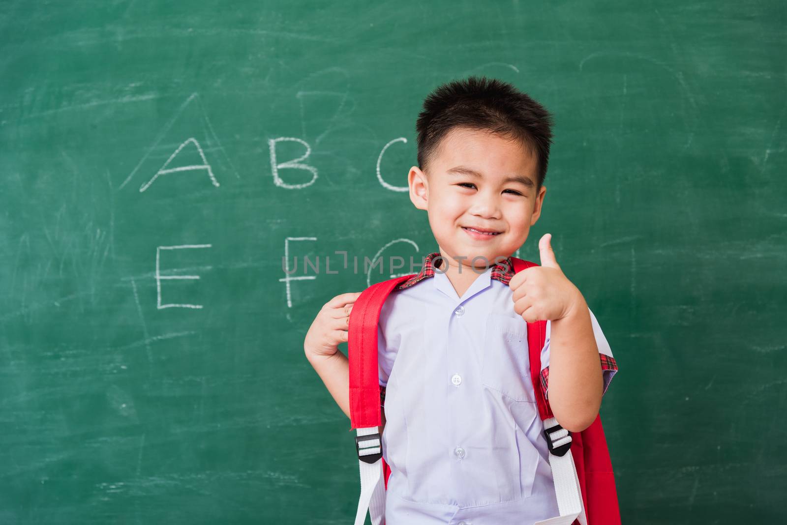 Back to School. Happy Asian funny cute little child boy kindergarten in student uniform with school bag smiling show finger thumb up on green school blackboard, First time to school education concept