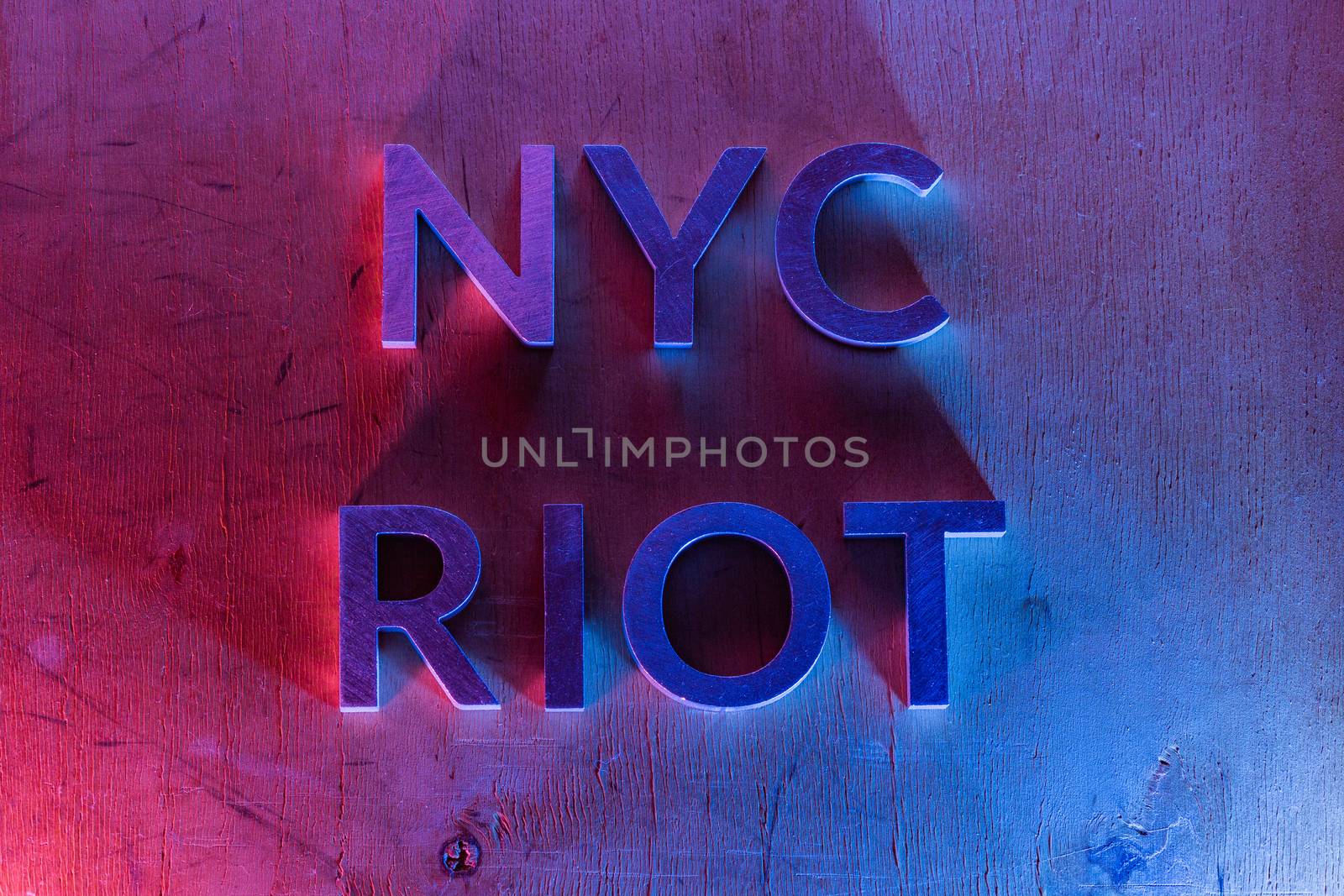 The words NYC riot laid with metal letters on flat plywood surface under red and blue police lights. Concept of night disorder in New York City.