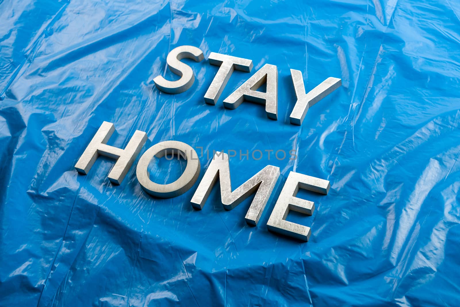 the words stay home laid with silver metal letters on crumpled blue plastic background in slanted diagonal perspective.