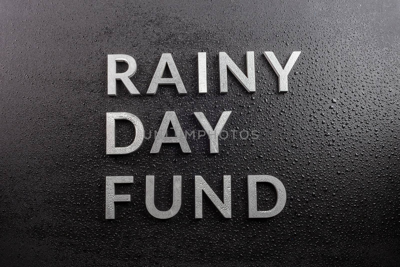 the words rainy day fund laid with silver metal letters on black background, covered with water drops, flat lay perspective, directly above view