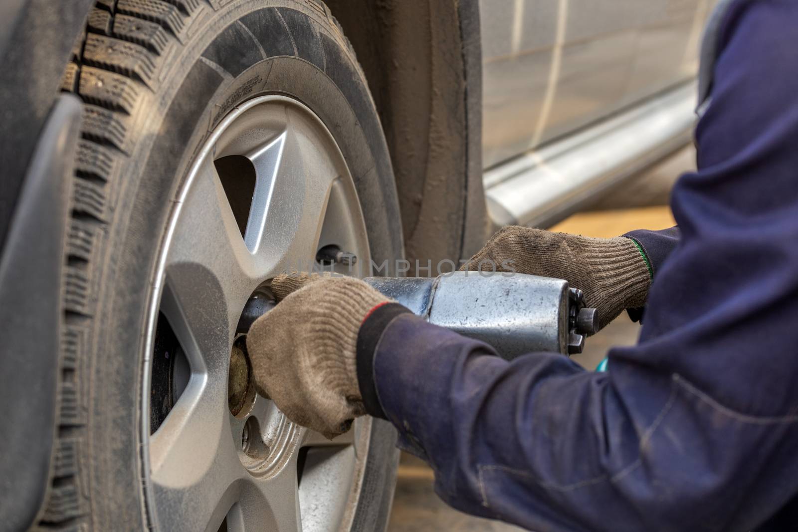 Hands of automotive mechanic unscrewing nuts with pneumatic impact wrench during car wheels season changing. Close-up view with selective focus and background blur.