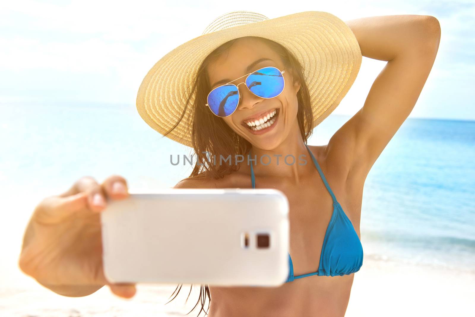 Selfie summer beach vacation travel girl taking self-portrait photo with phone on sun tan holiday. Woman wearing beach hat, mrror sunglasses, blue swimsuit.