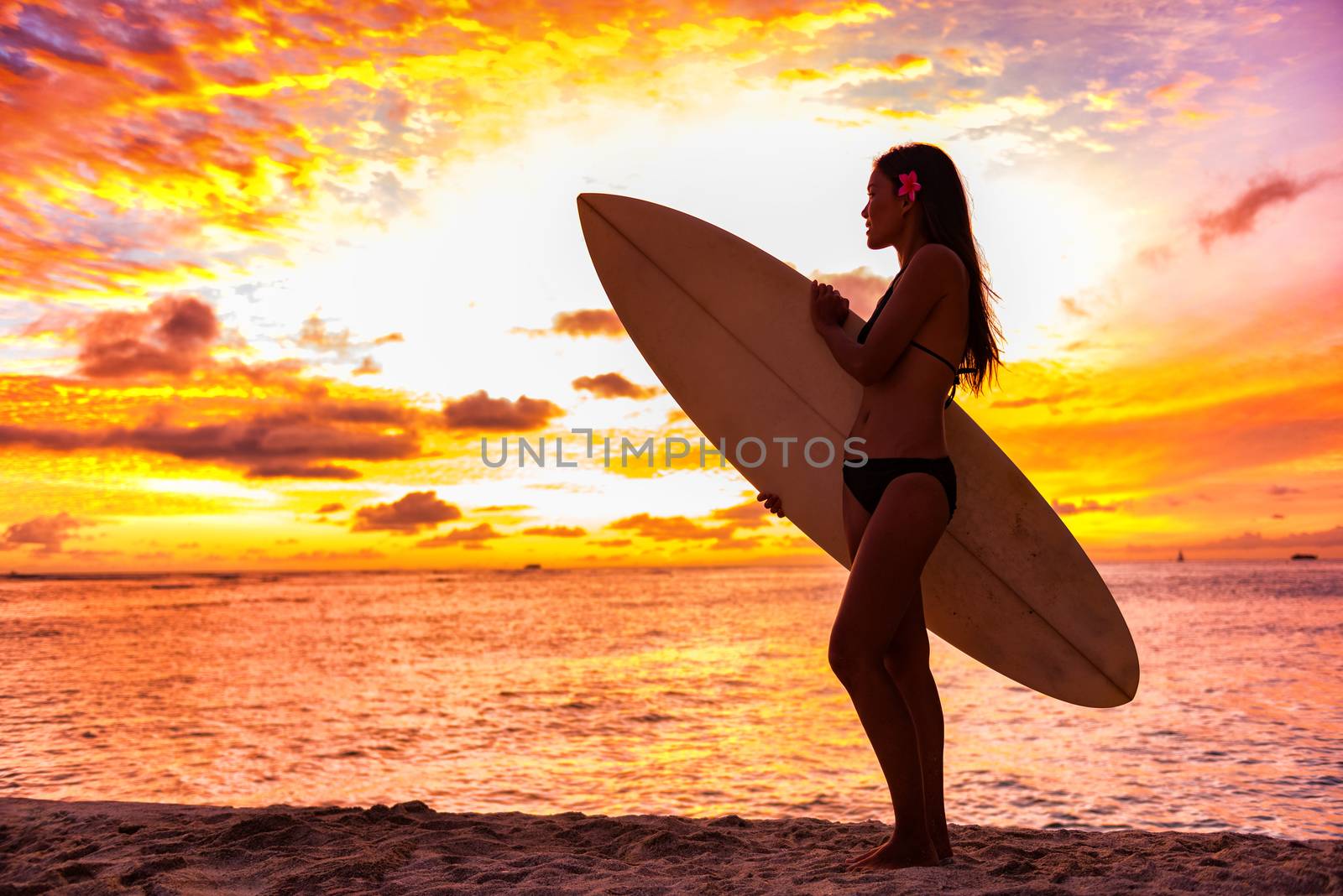 Surfer bikini girl on Hawaii beach holding surf board watching ocean waves at sunset. Silhouette of Asian sport woman over landscape, sky and clouds background. Summer vacation lifestyle.