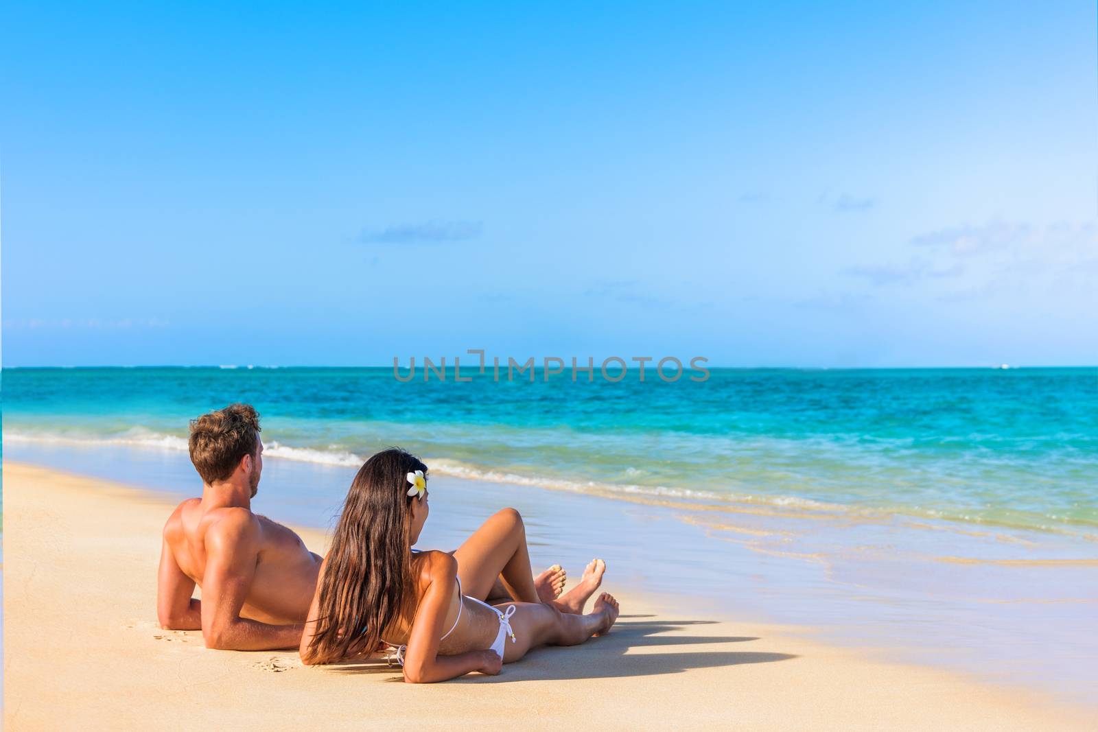 Couple on beach travel honeymoon vacation lying down sunbathing relaxing on luxury holiday in idyllic destination. Young tourists in love by Maridav