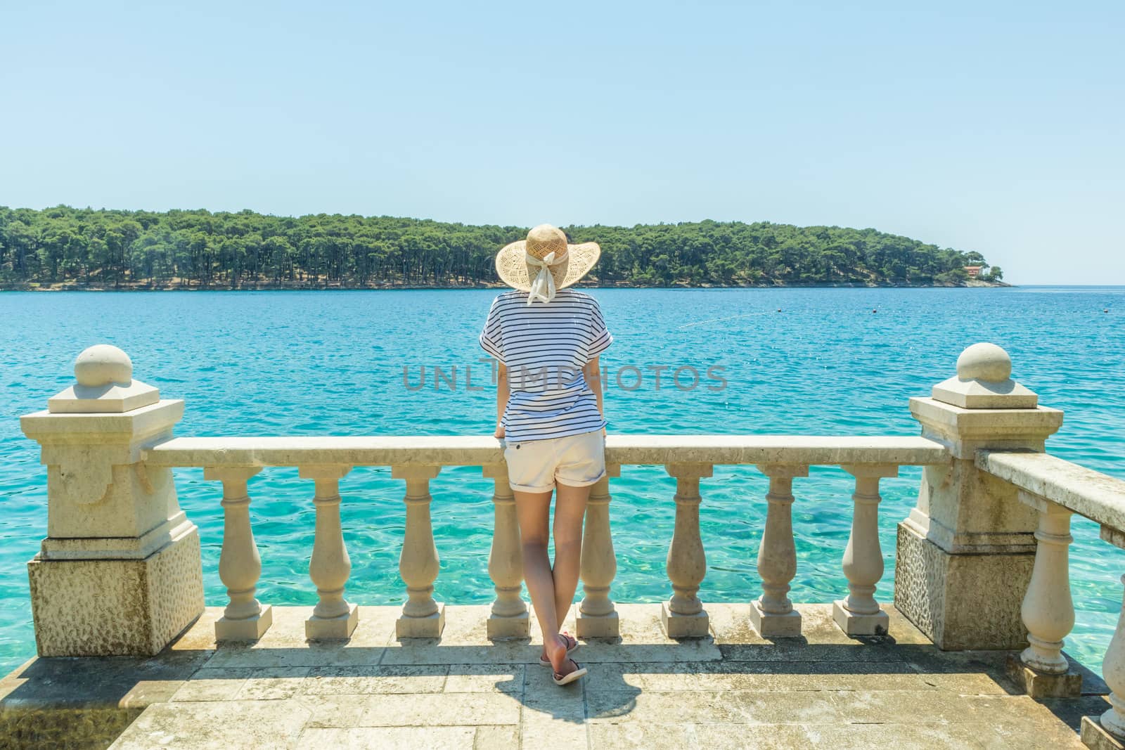 Rear view of woman wearing straw summer hat ,leaning against elegant old stone fence of coastal villa, relaxing while looking at blue Adriatic sea, on Losinj island Croatia. by kasto