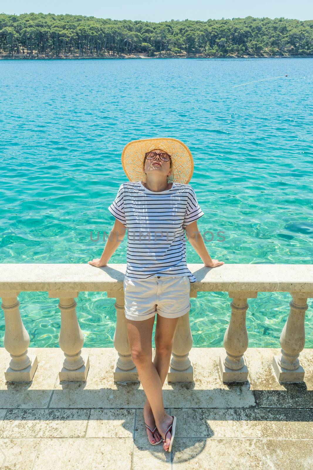 Rear view of woman wearing straw summer hat ,leaning against elegant old stone fence of coastal villa, relaxing while looking at blue Adriatic sea, on Losinj island Croatia. by kasto