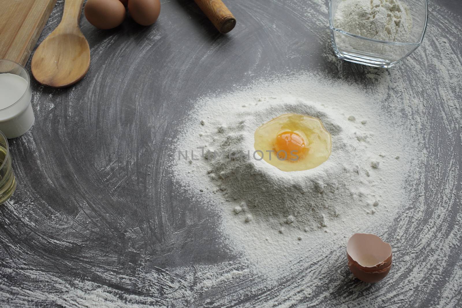 Broken chicken egg in a pile of flour, olive oil, milk, kitchen tool, gray table by selinsmo