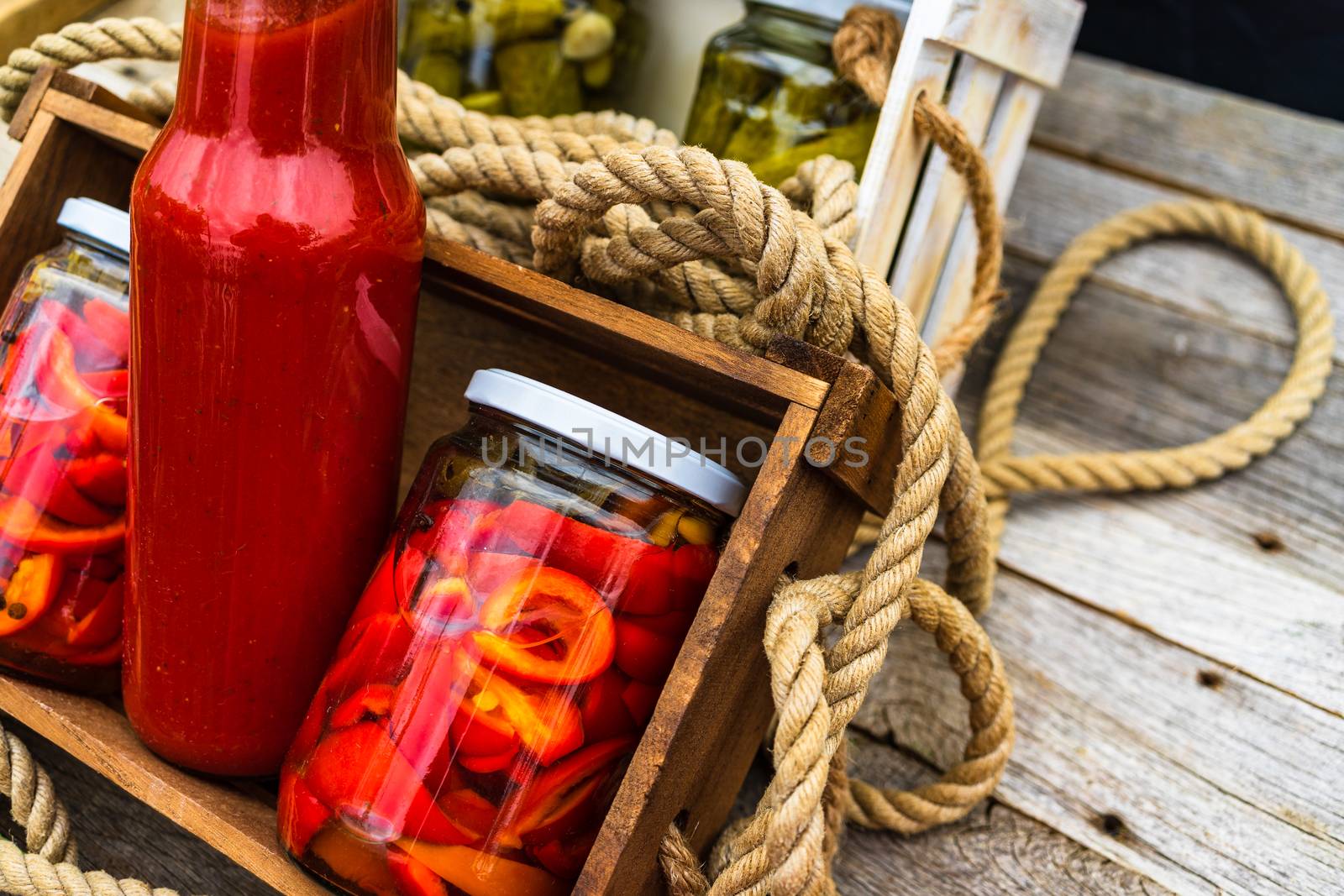 Wooden crate with bottles with tomatoes sauce and glass jars with pickled red bell peppers isolated in a rustic composition. Jars with variety of pickled vegetables preserved food concept.