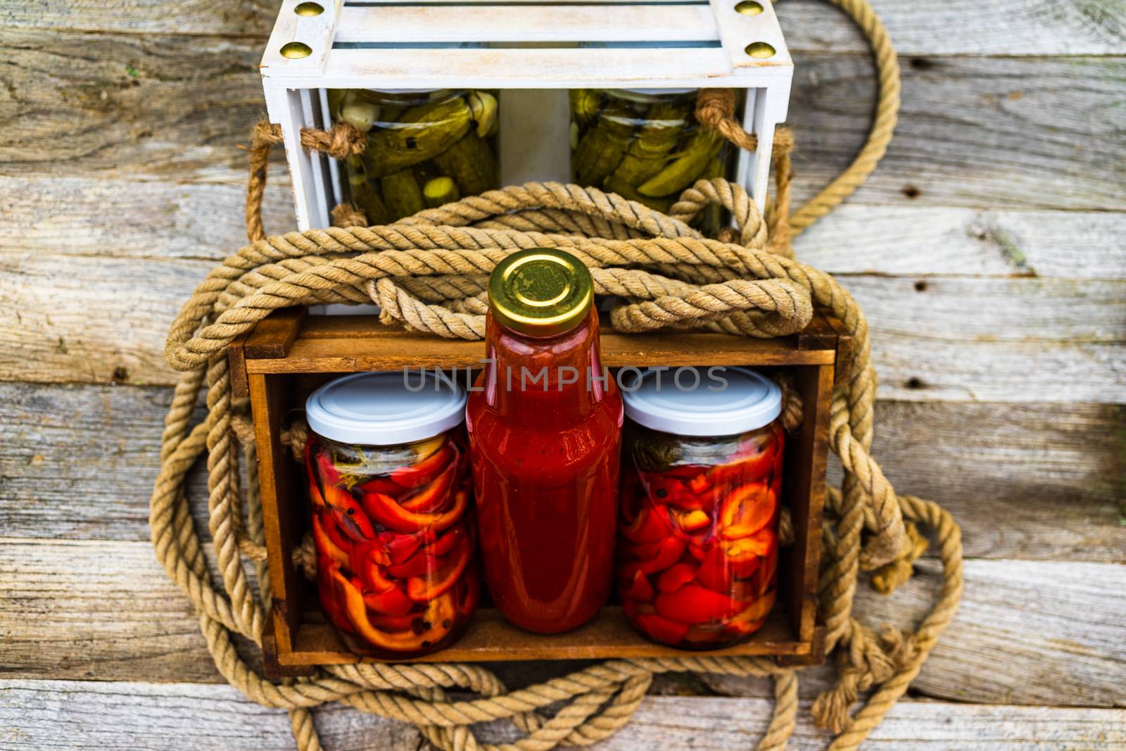 Wooden crate with bottles with tomatoes sauce and glass jars with pickled red bell peppers isolated in a rustic composition. Jars with variety of pickled vegetables preserved food concept.