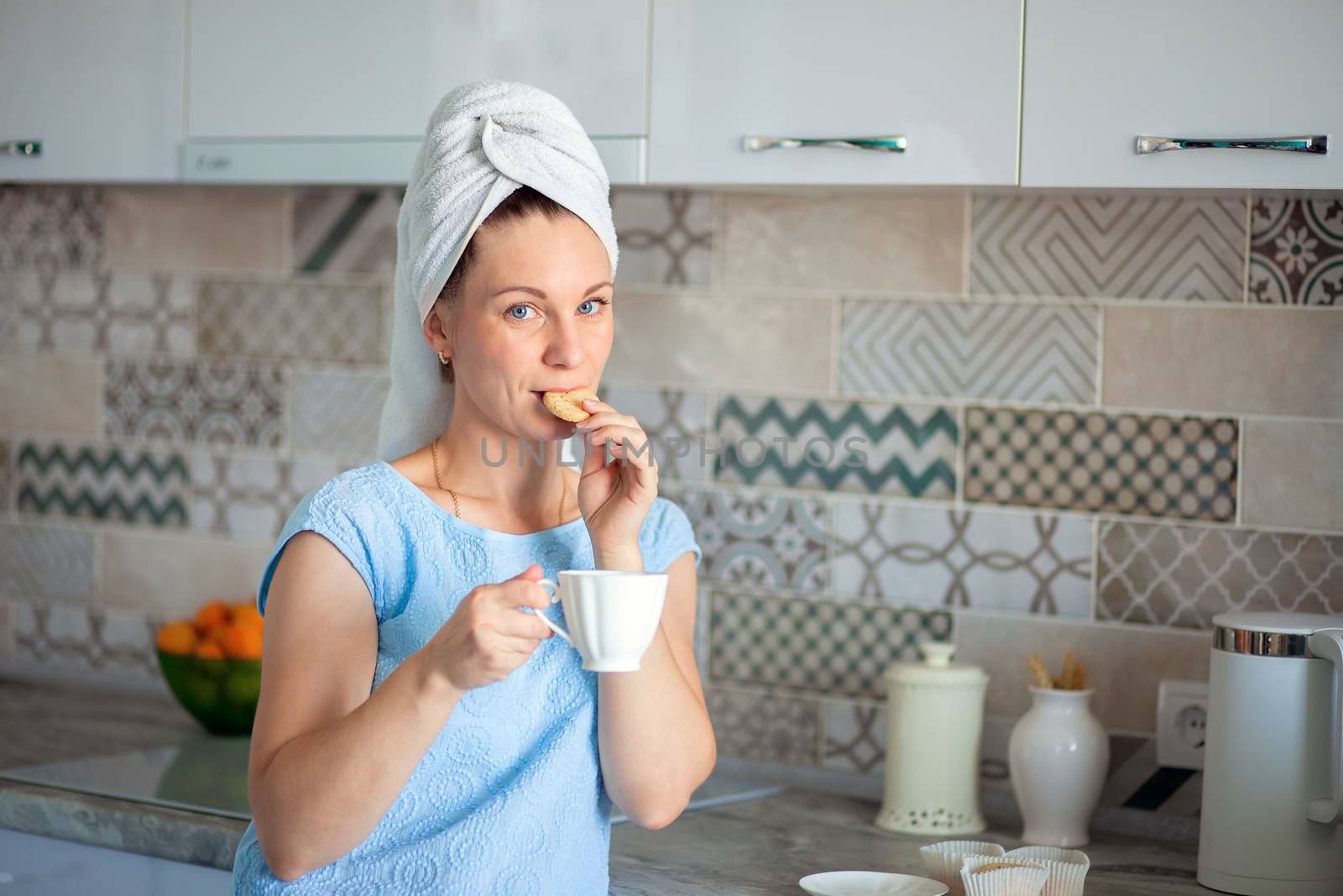 A satisfied woman washed her hair in the morning and, with a towel on her head, drinks coffee and eats cookies. Morning toilet and breakfast