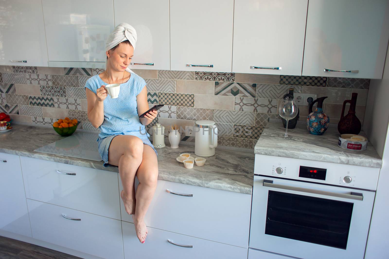 A girl with a towel on her head in her kitchen drinks coffee and looks at the phone