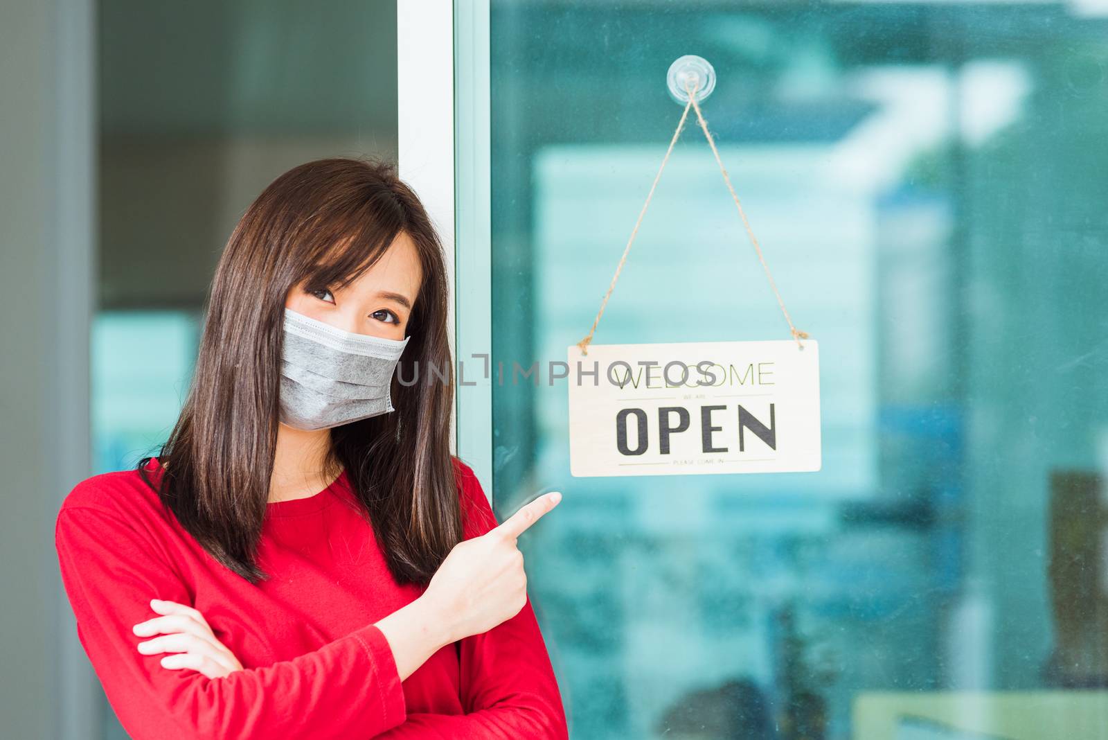 New normal Asian young woman wear face mask protect glad point finger to notice sign wood board label "WELCOME OPEN" hang through glass door front shop, Business turn open after coronavirus pandemic