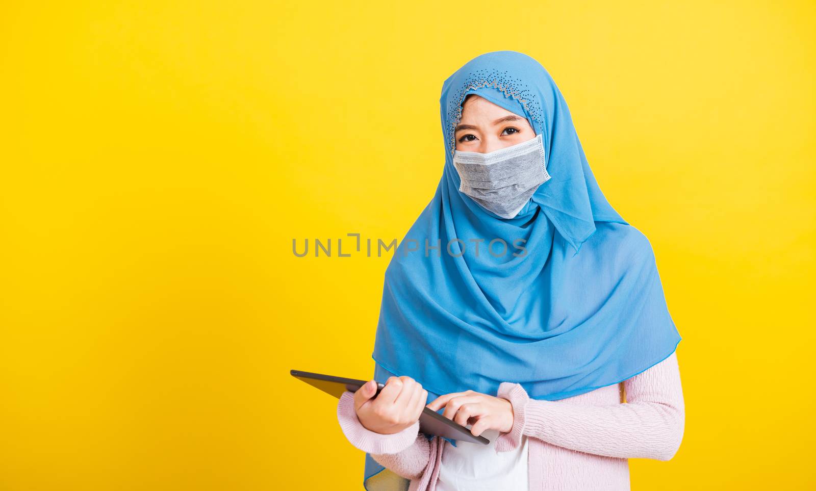 Asian Muslim Arab, Portrait of happy beautiful young woman Islam religious wear veil hijab and face mask protect she quarantines disease coronavirus touch on tablet screen isolated yellow background