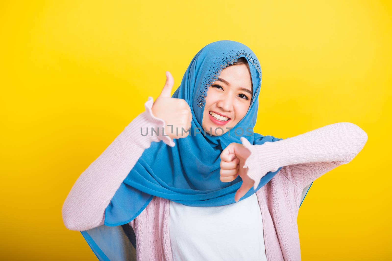 Asian Muslim Arab, Portrait of happy beautiful young woman Islam religious wear veil hijab funny smile she showing thumbs up and thumbs down difficult choose isolated yellow background