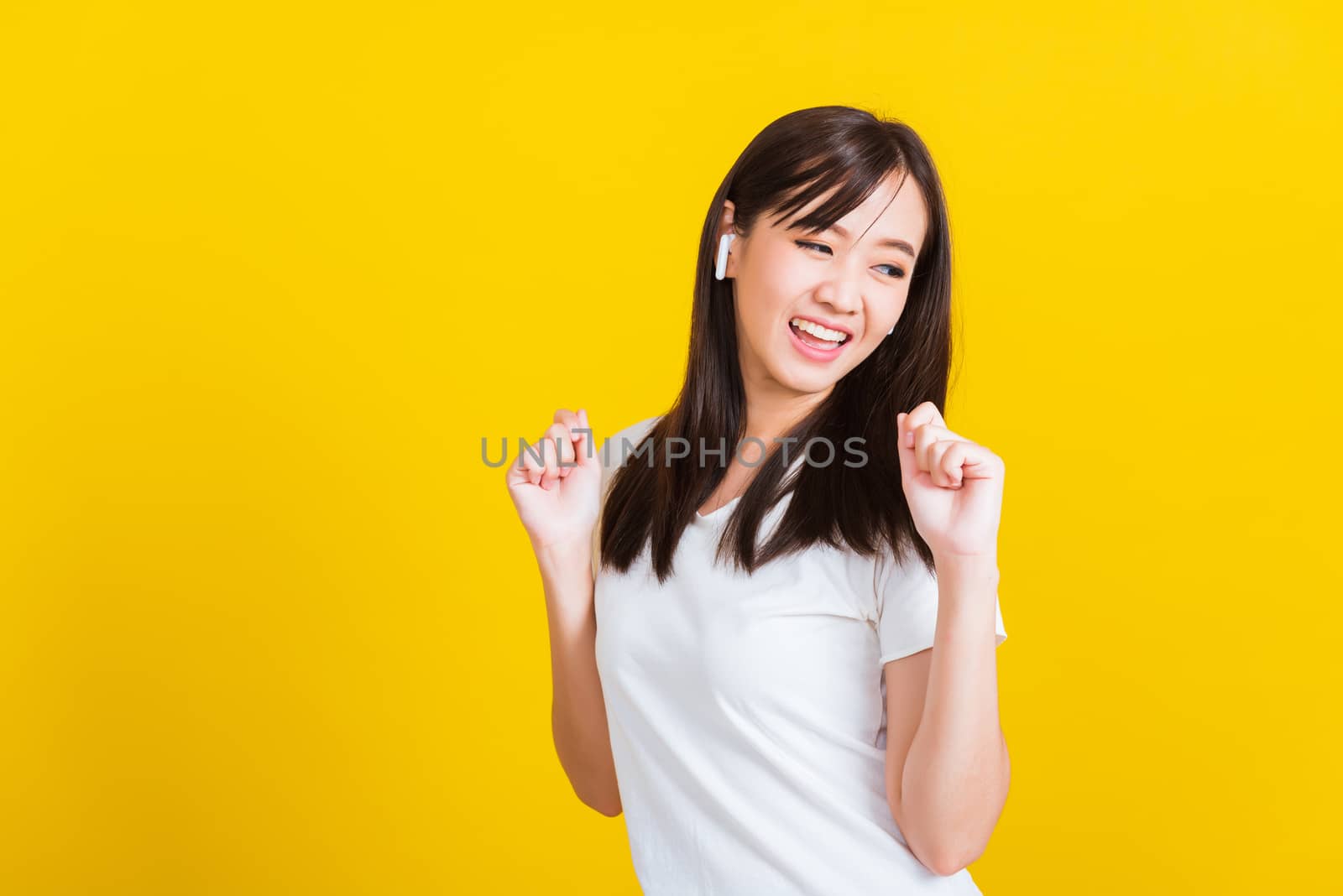 Portrait Asian of a happy beautiful young woman wear wireless earphone listening to music from smartphone having fun and dancing to sounds of favorite music studio shot isolated on yellow background