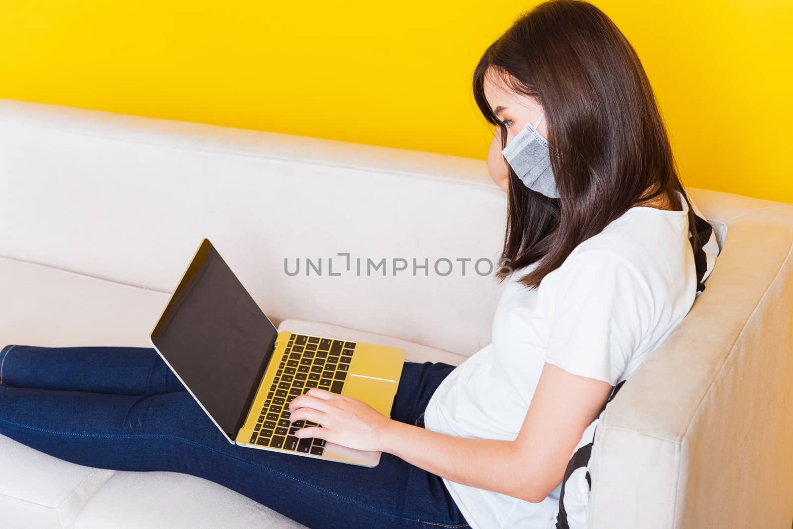 Portrait Asian of beautiful young woman sitting on sofa wearing medical face mask protective she work from home with laptop computer during Coronavirus studio shot isolated on yellow background