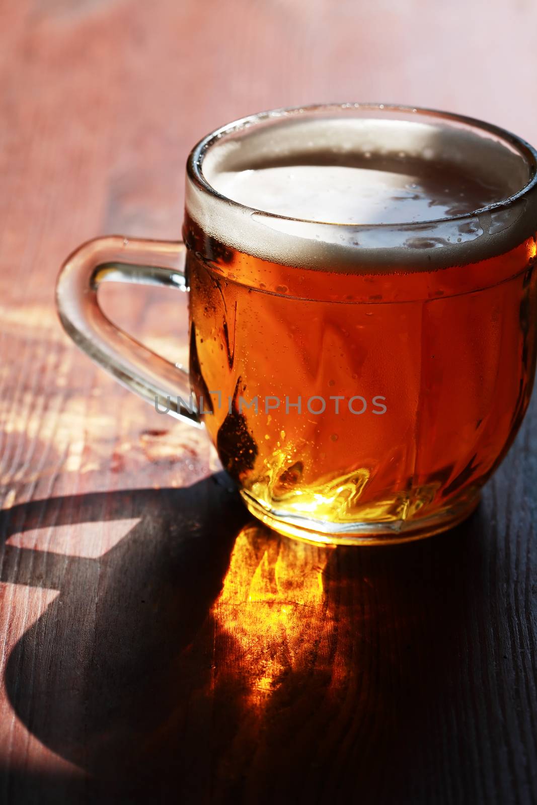 Mug of beer with foam on wooden table under sunlight