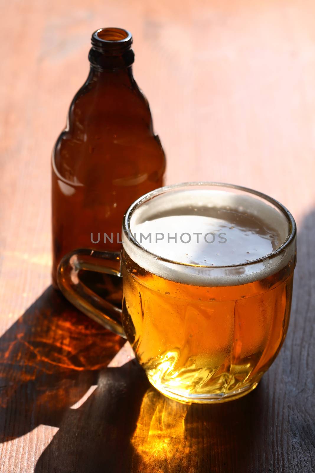Mug of beer with foam on wooden table under sunlight