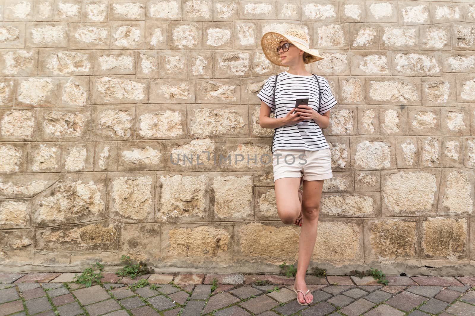 Beautiful young female tourist woman standing in front of old textured stone wall at old Mediterranean town, smiling, holding, smart phone to network on vacationes. Copy space.