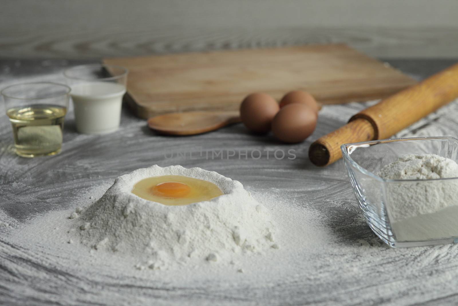 Broken chicken egg in a pile of flour, olive oil, milk, kitchen tool on gray table background. Products for baking bakery products. Cutting board, rolling pin, flour sieve, wooden spoon. bread or cake