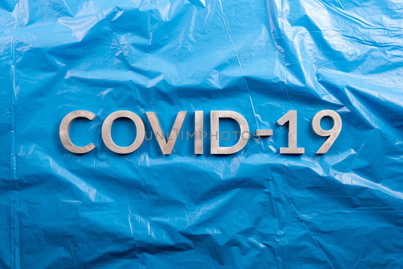 The word COVID-19 laid with aluminium letters on crumpled blue plastic film background in flat lay composition, directly above view.