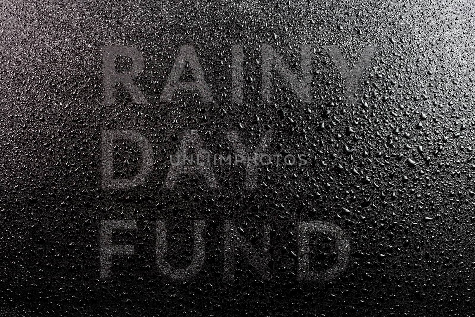 the dry words rainy day fund covered from water drops on black surface, unusual implementation concept in flat lay perspective.