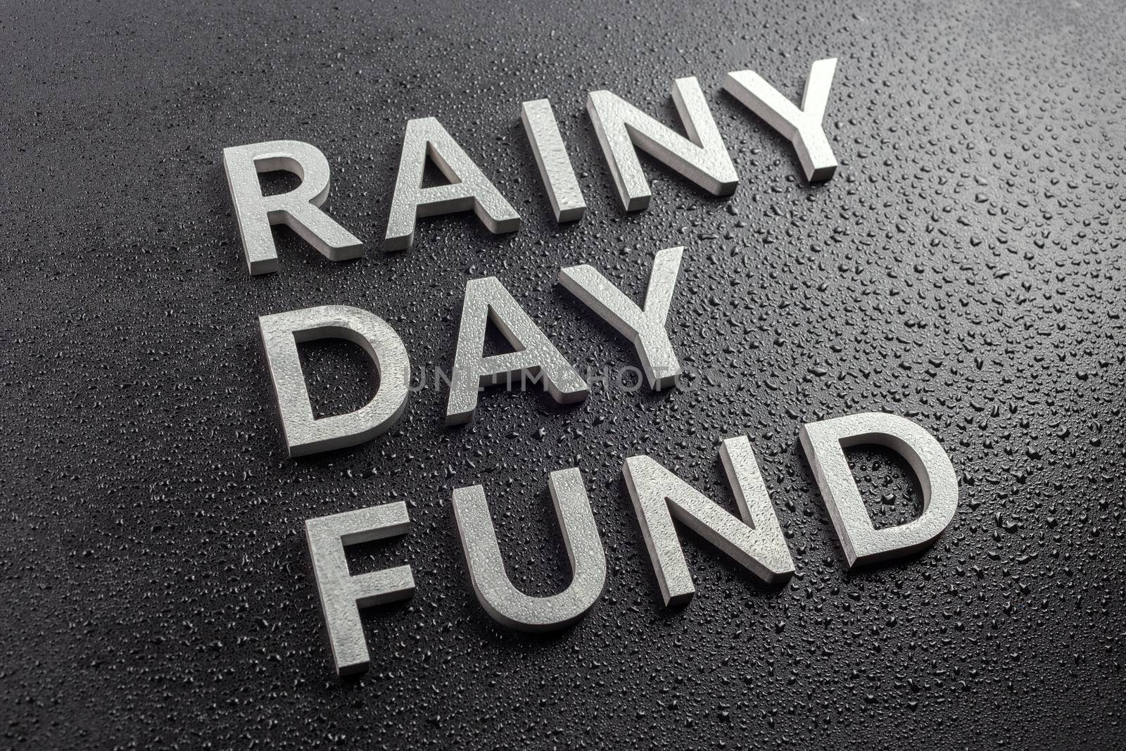 the words rainy day fund laid with silver metal letters on black background, all covered with water drops, slanted diagonal perspective with selective focus and blur