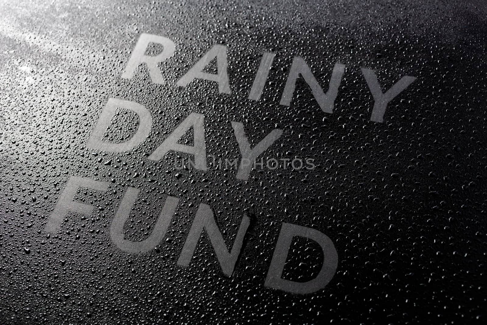 the dry words rainy day fund covered from water drops on black surface, unusual implementation concept with slanted diagonal perspective with selective focus