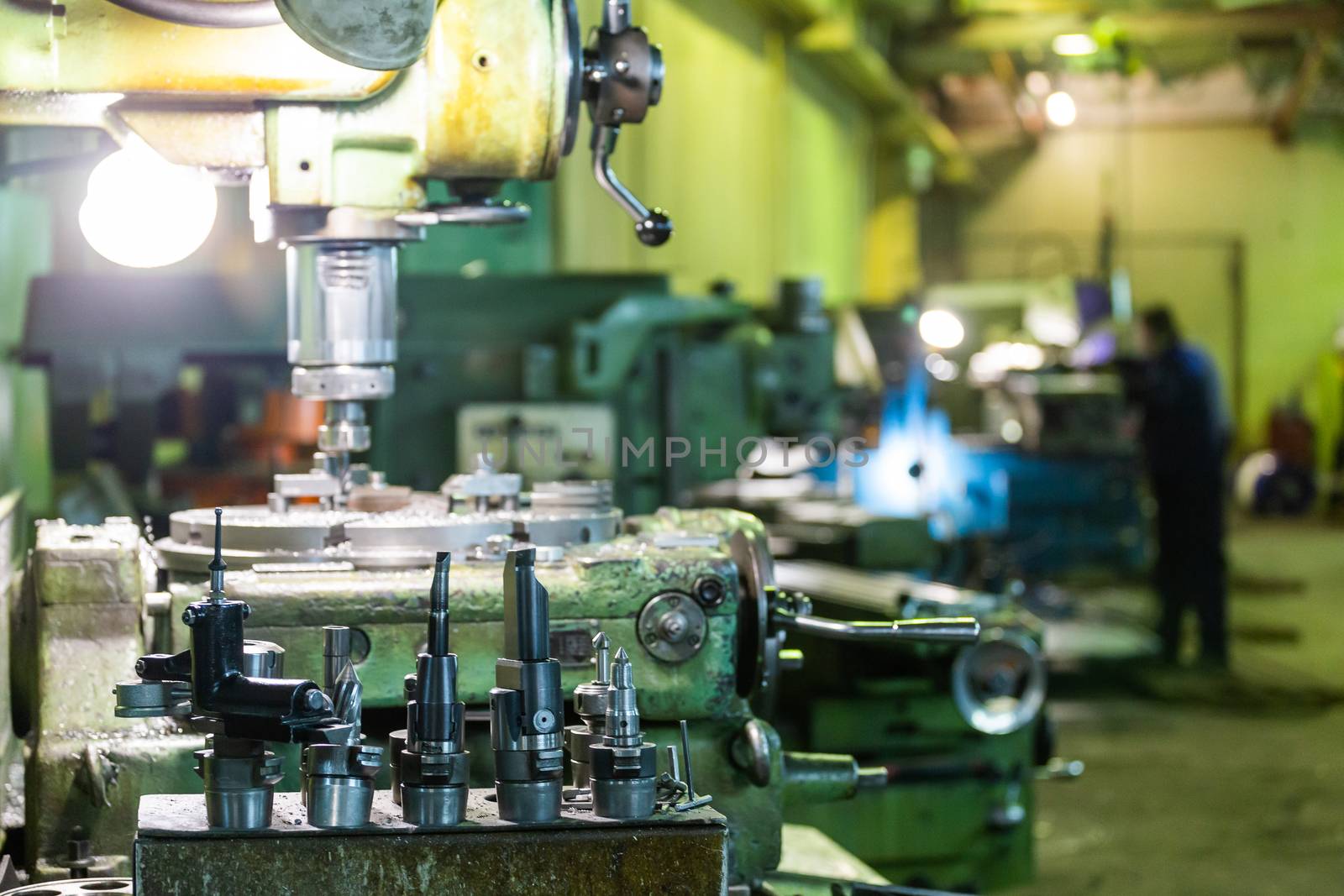 A set of metalworking tools in front of manual vertical milling and boring oldschool machine indoors with working people in blurry background,