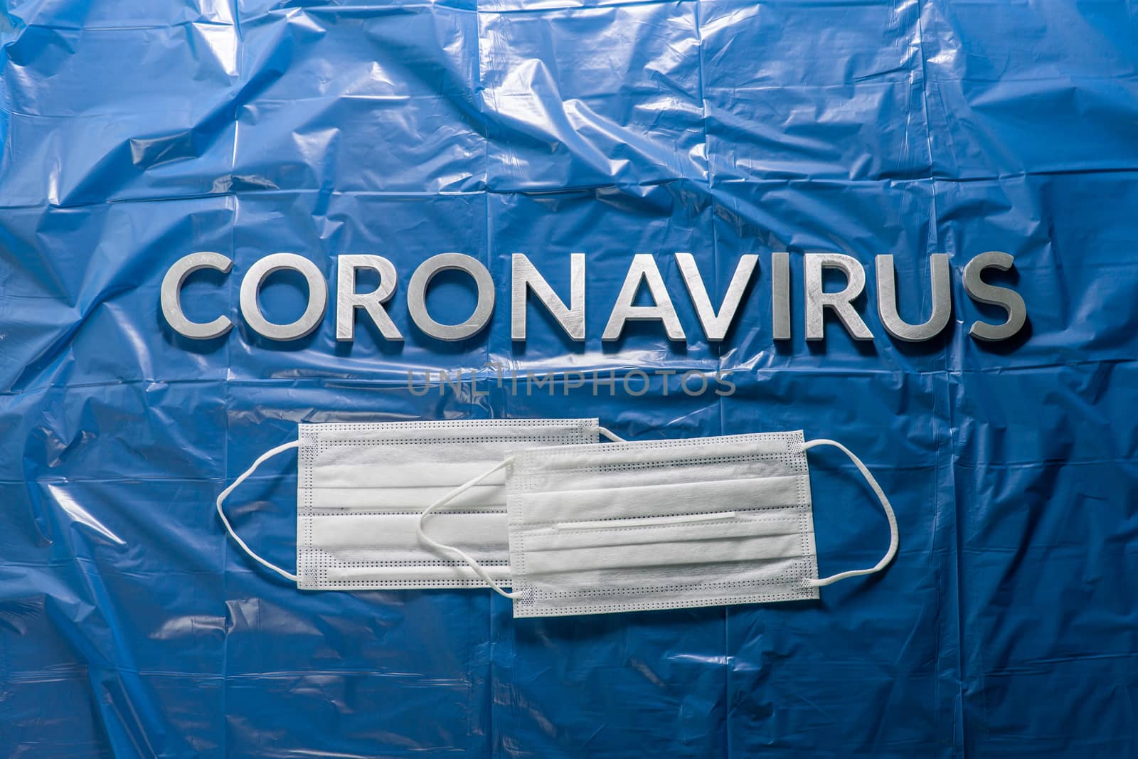 the word coronavirus laid with silver letters on crumpled blue plastic film with white medical facial masks - flat lay, centered composition and dramatic light