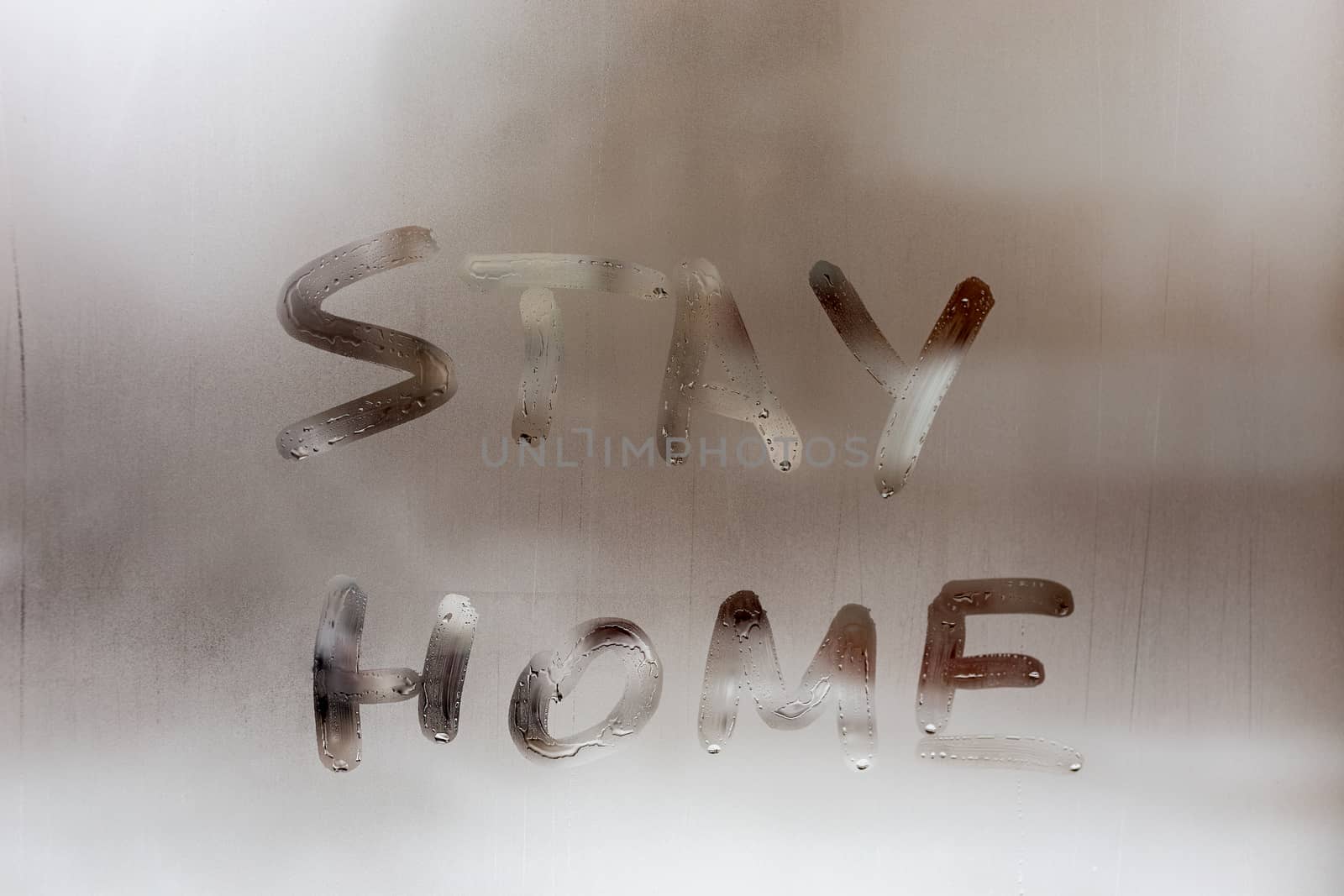 the words stay at home handwritten on wet window glass at daytime - close-up full frame picture with selective focus and background blur