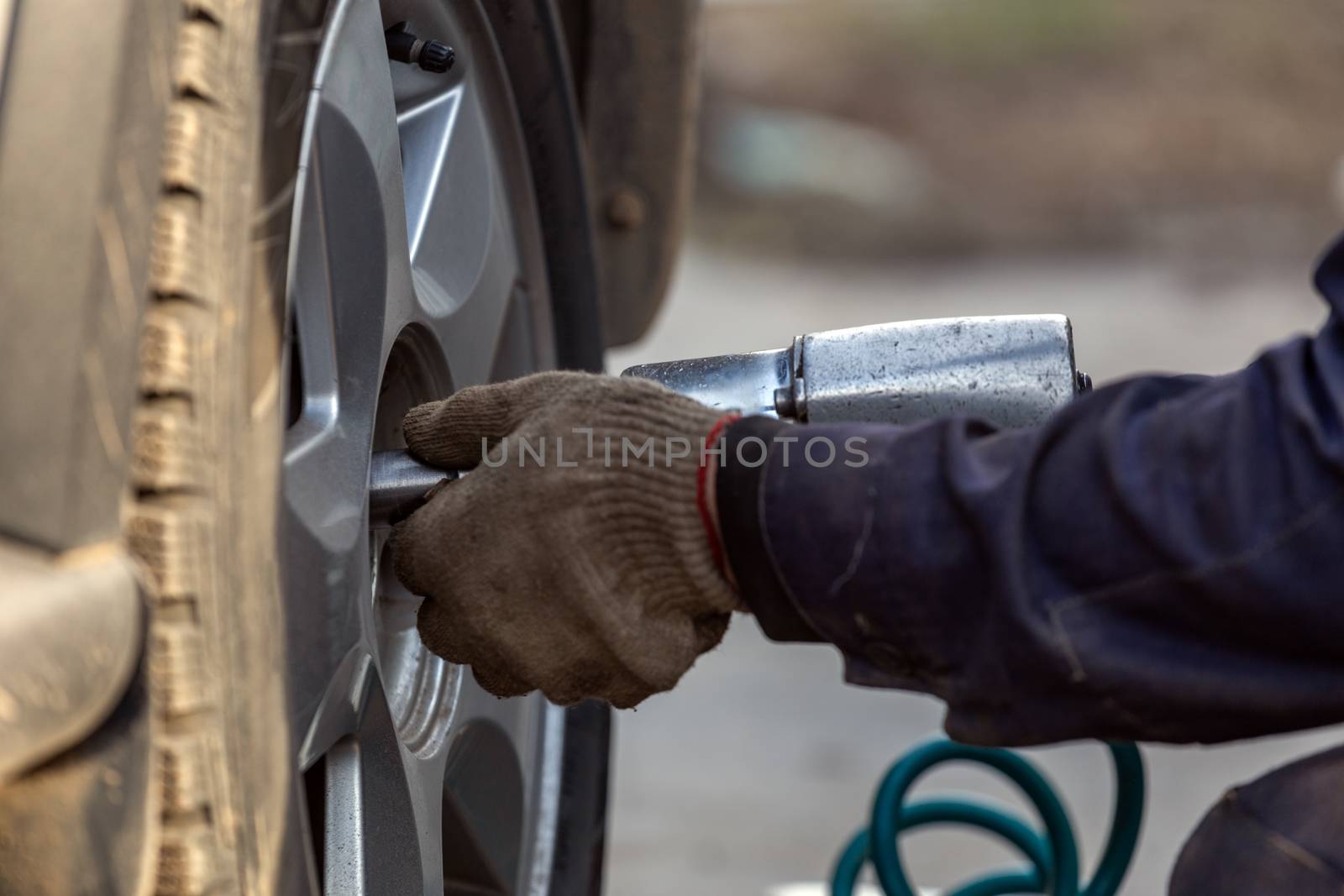 Hands of automotive mechanic unscrewing nuts with pneumatic impact wrench during car wheels season changing. Close-up view with selective focus and background blur.