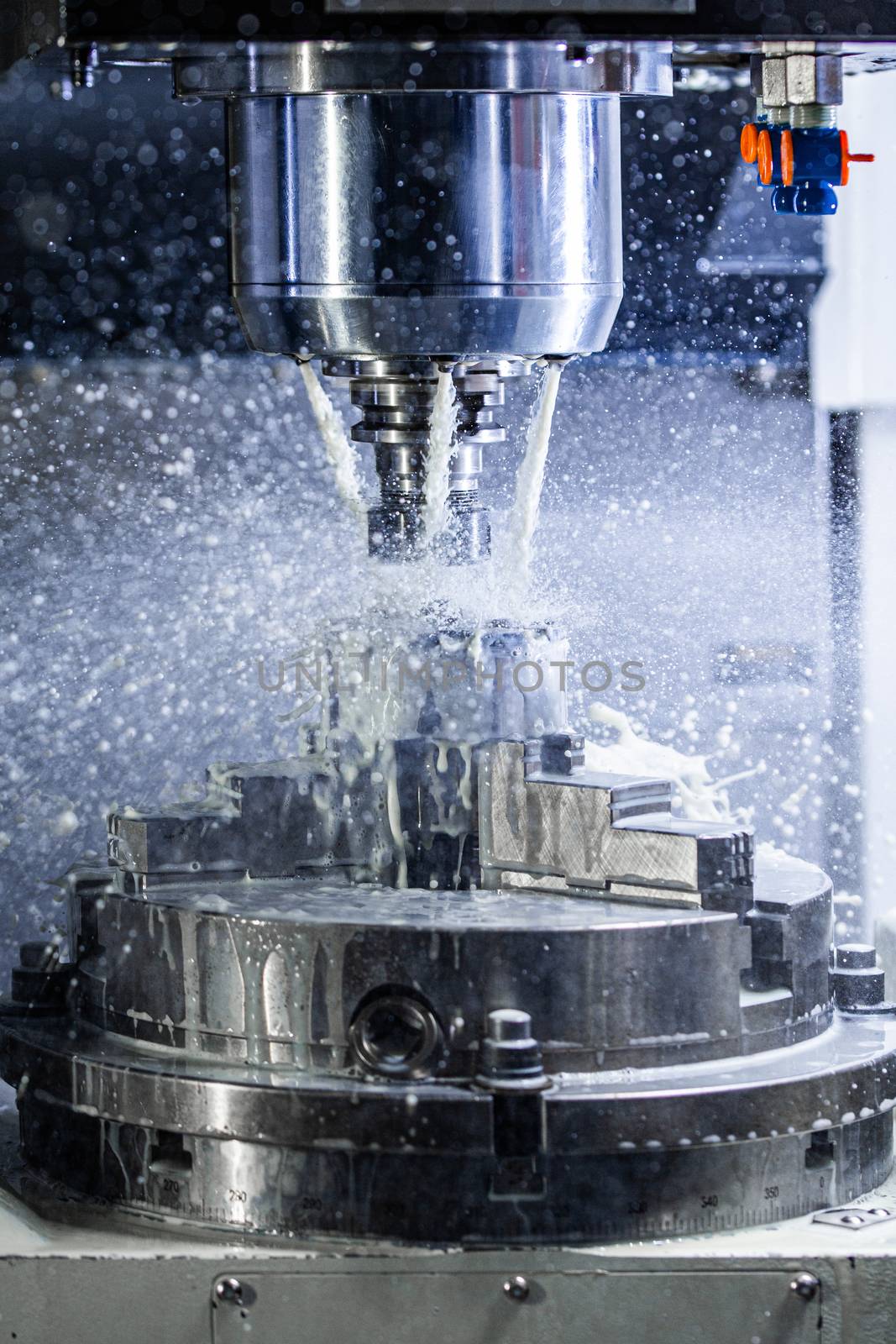 Vertical photo of industrial wet milling process in 5-axis cnc machine with coolant flow under pressure and freezed splashes.