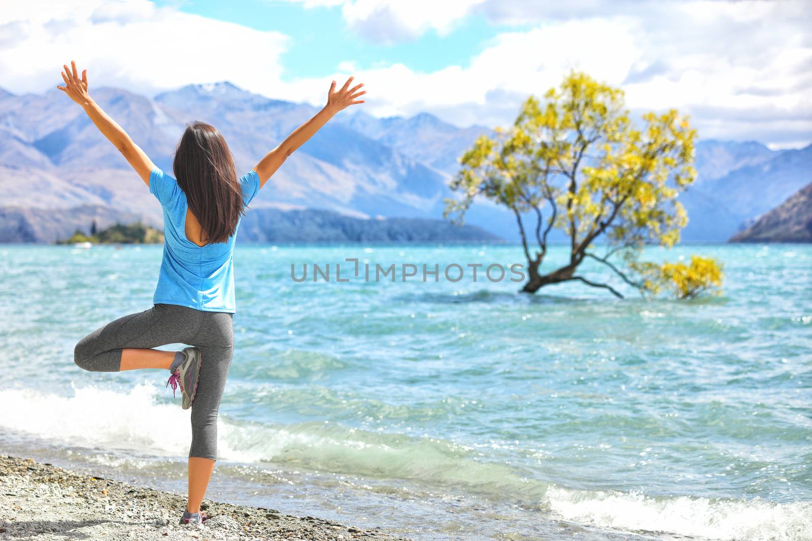 New Zealand morning yoga girl doing tree pose variation doing yoga practice with open arms at Wanaka lake by the lone tree, tourist travel popular attraction in New Zealand. Beach nature landscape by Maridav