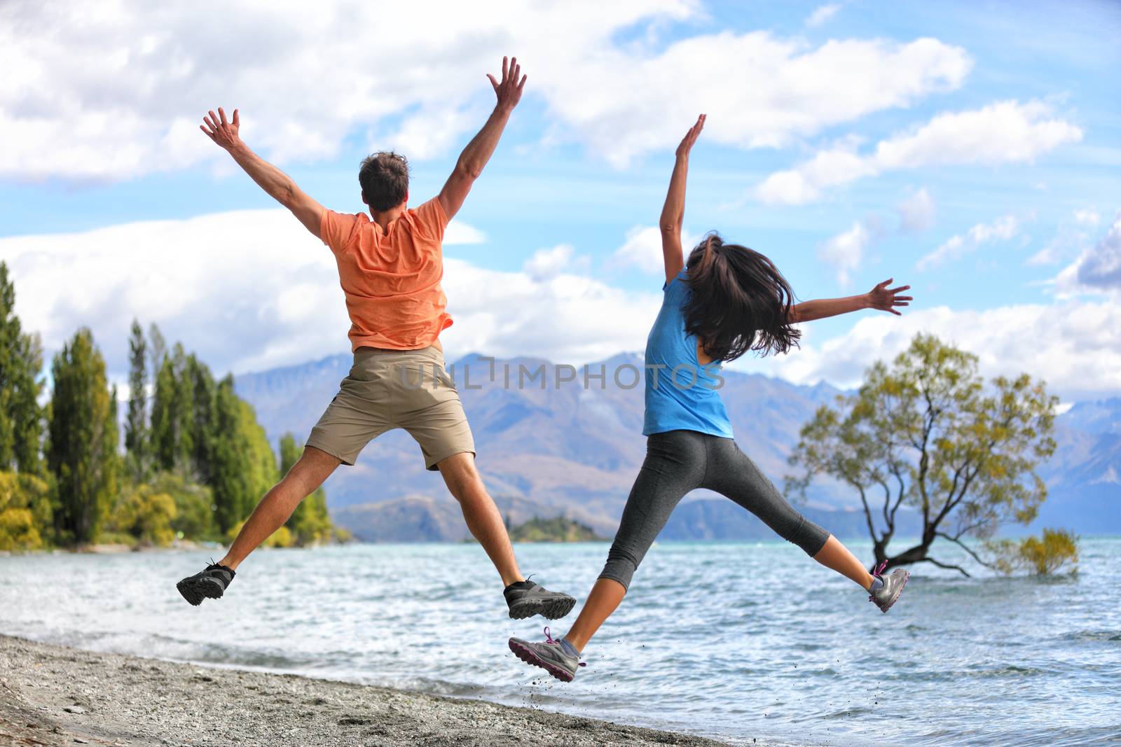 New Zealand tourists couple jumping of happiness at Wanaka Lone . Happy young people doing funny jump of joy sightseeing traveling in summer adventure destination. Woman and man.