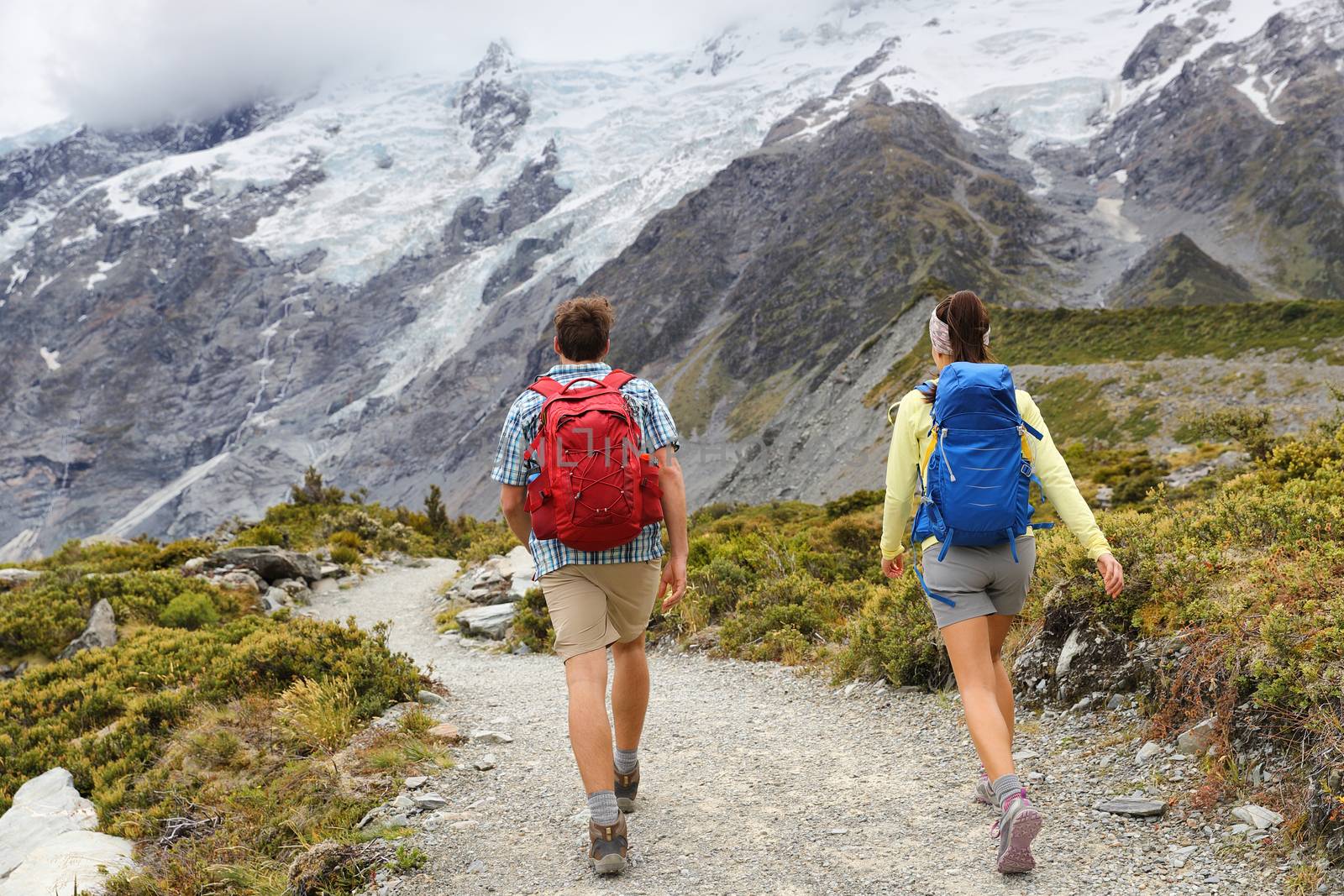 New Zealand travel tramping hikers walking on trail of Hooker Valley Track in New Zealand Aoraki/Mt Cook. Couple tourists hiking in mountains on popular tourist destination. Kiwi trampers with bags.