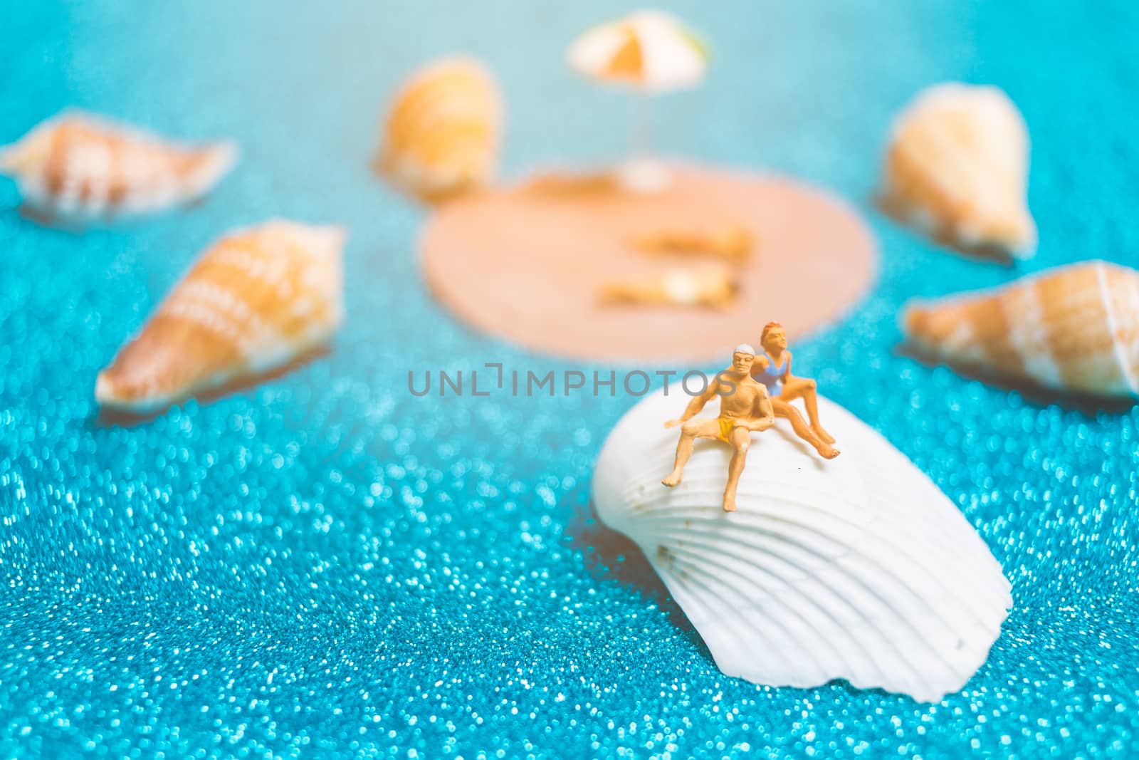 Miniature people wearing swimsuit relaxing on a seashell with blue glitter background