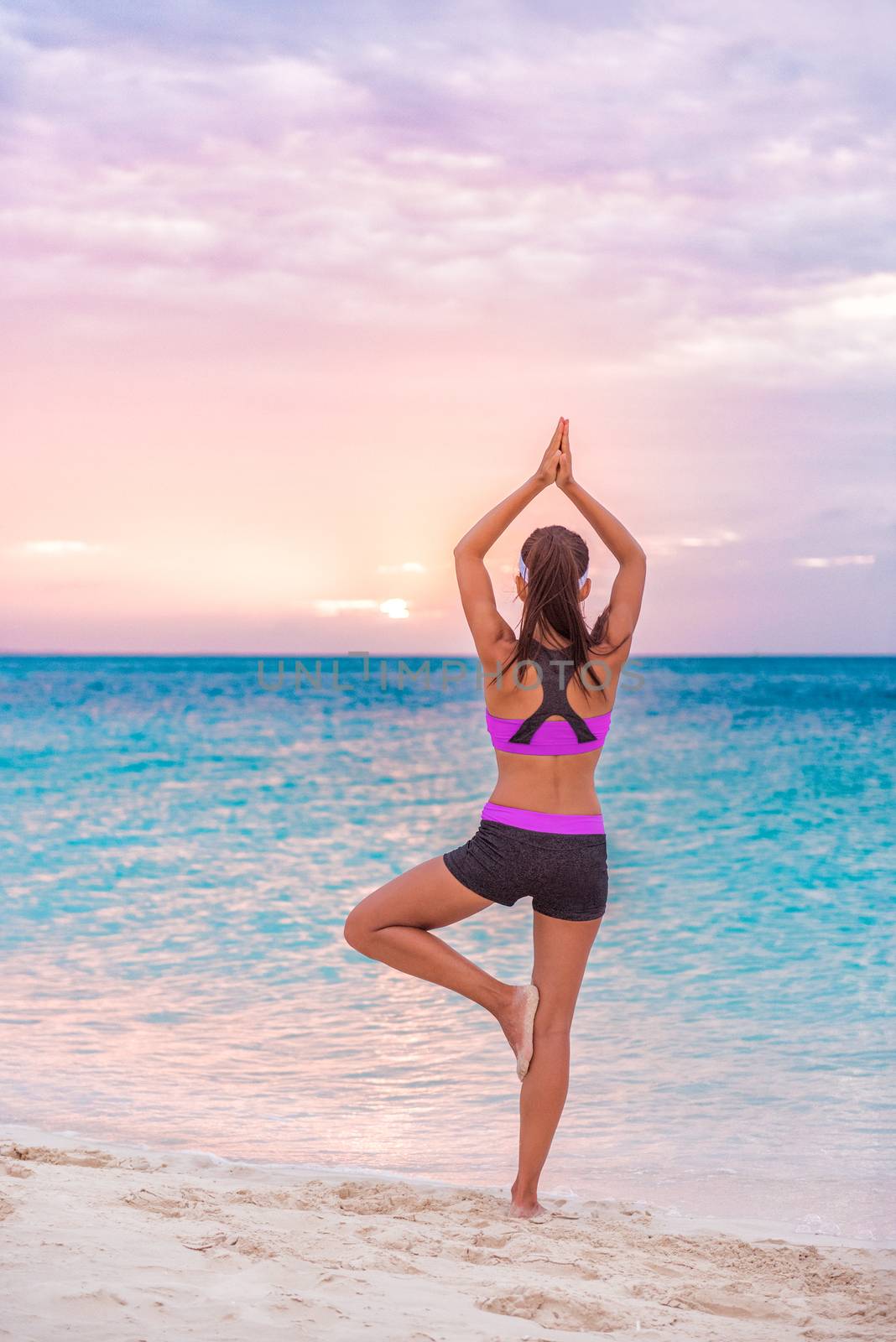 Yoga wellness class on beach at sunset. Girl practicing meditation standing on one leg training balance barefoot on sand enjoying sun and ocean view by Maridav