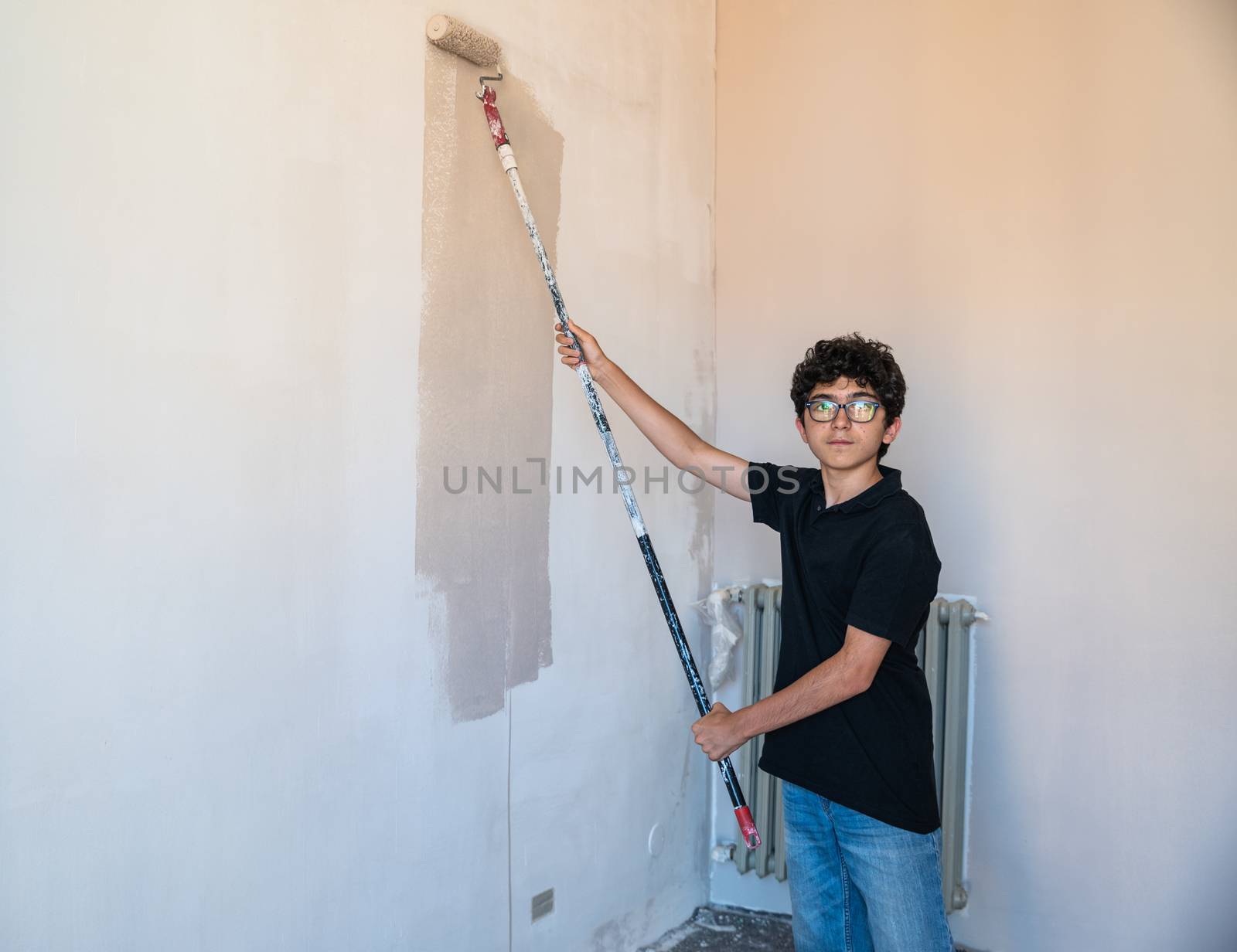Renovate the walls of the house by painting them: the Caucasian boy with glasses passes the gray impregnated roller on the wall. A little color drips down the wall.