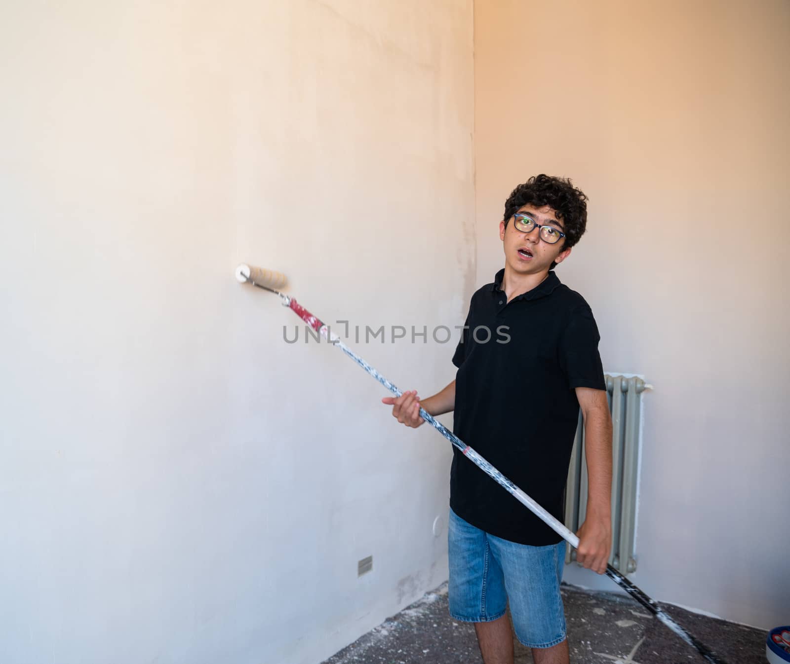 Renovate the walls of the house by painting them: the Caucasian boy with glasses passes the roller on the wall. He's not happy about doing this job!