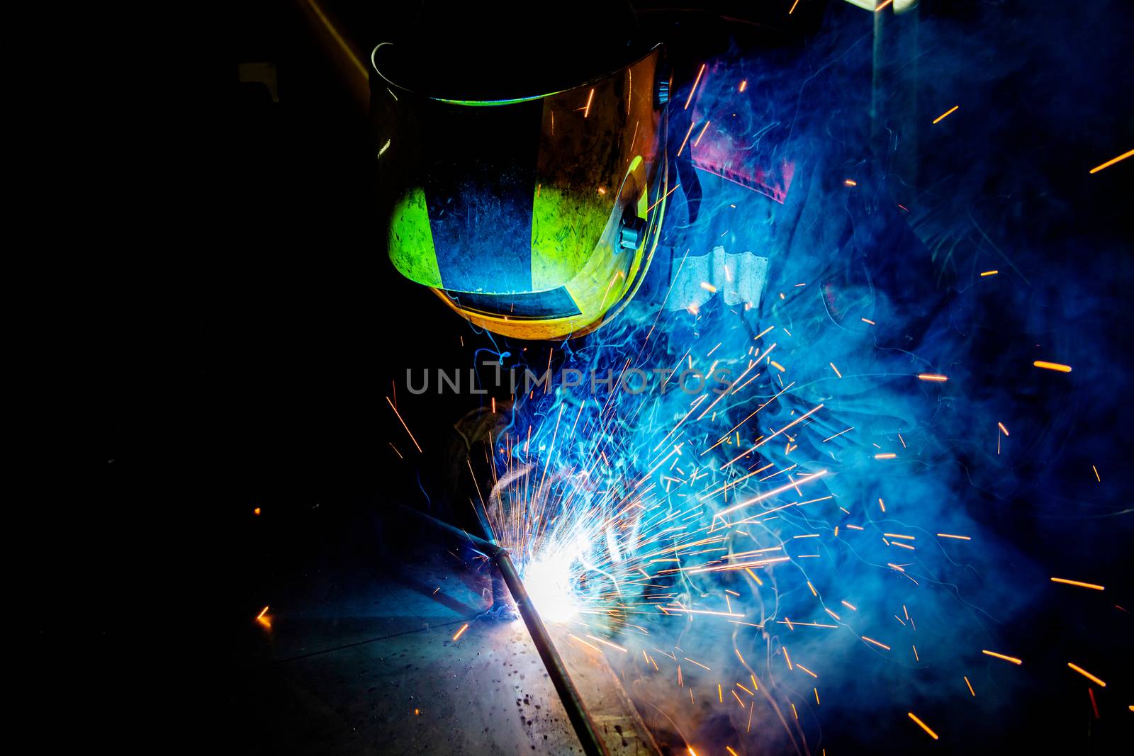 A process of manual operational semi-automatic electric welding with sparks, professional industrial worker in black-yellow protective mask at work.. High contrast dark close-up scene. MIG - metal inert gas - type of welding.