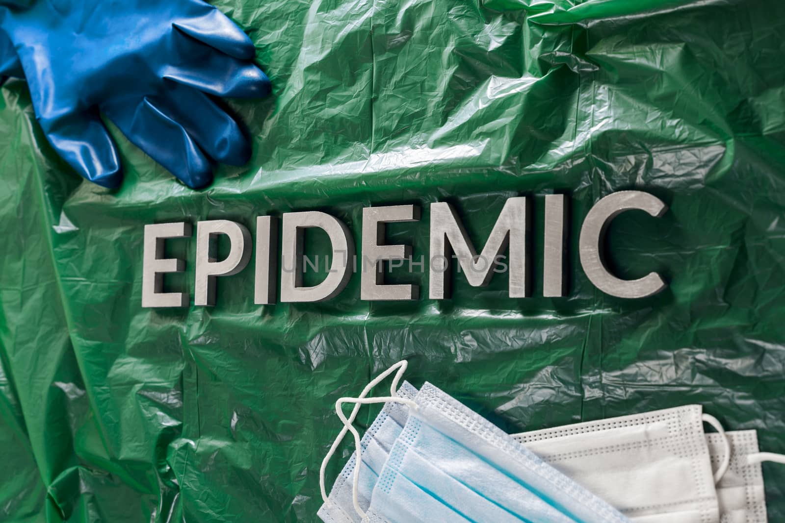 the word epidemic laid by metal letters in crumpled green plastic background with face masks and blue protective glove.