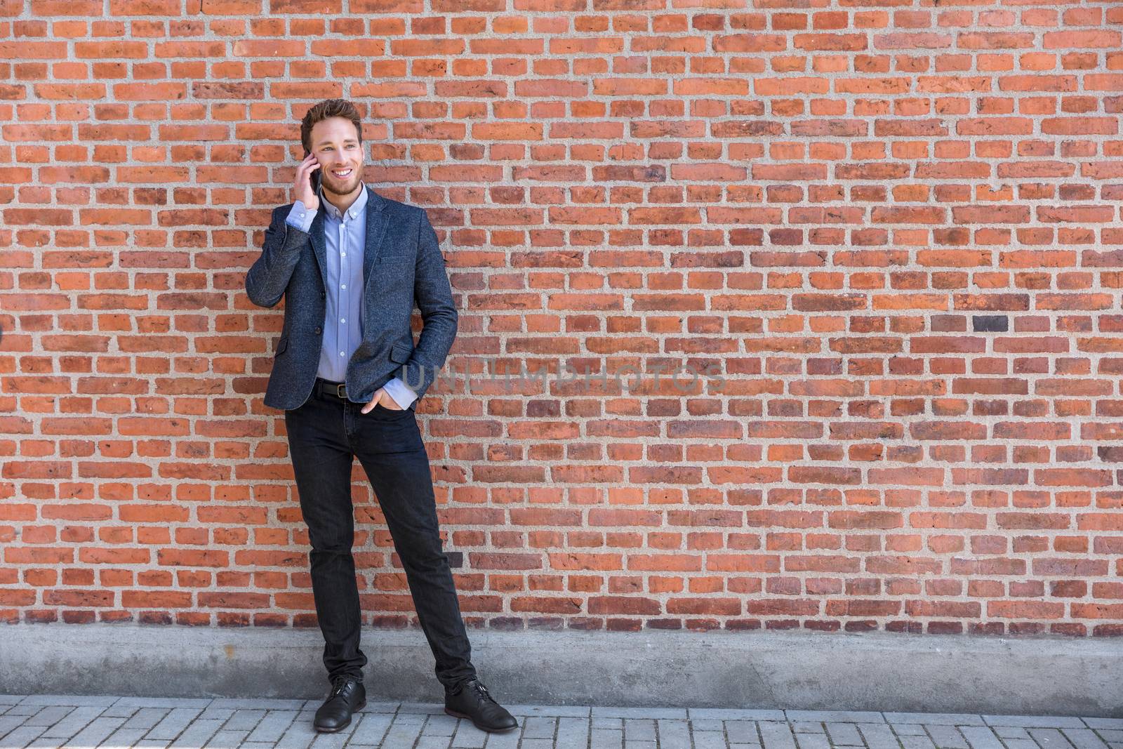 Man talking business on phone holding smartphone in city street in smart casual wear standing against brick wall urban background. Happy caucasian businessman by Maridav