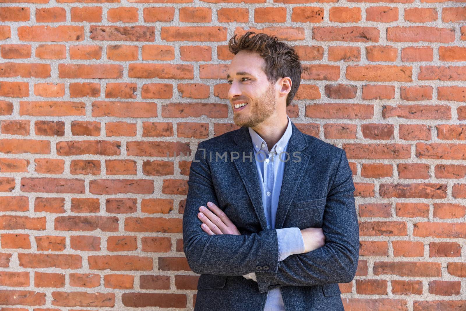Confident business entrepreneur man young businessman looking to the side portrait against city office brick wall background. Smiling caucasian male professional in smart casual jacket by Maridav