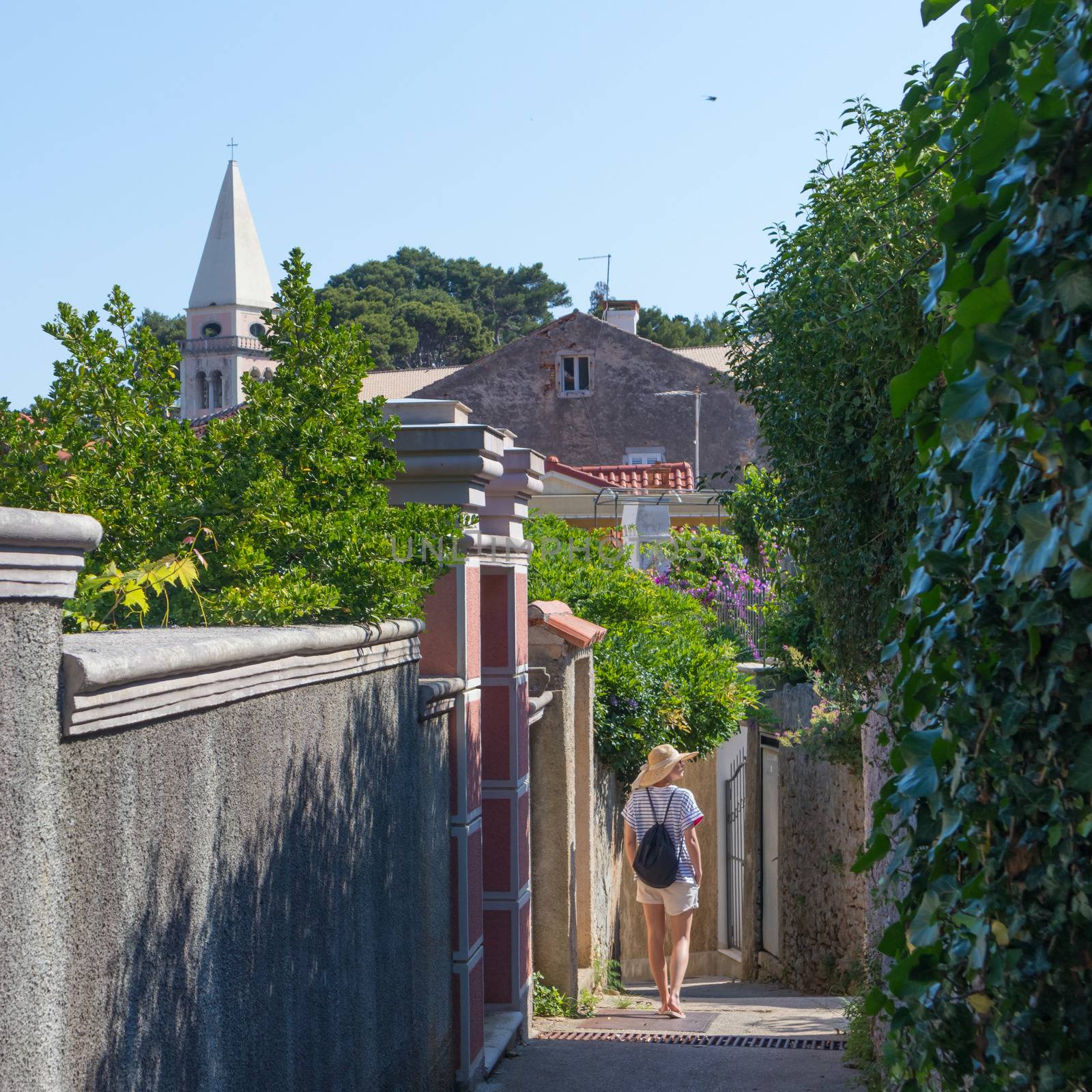 Rear view of beautiful blonde young female traveler wearing straw sun hat sightseeing and enjoying summer vacation in an old traditional costal town of Veli Losinj, Adriatic cost, Croatia by kasto