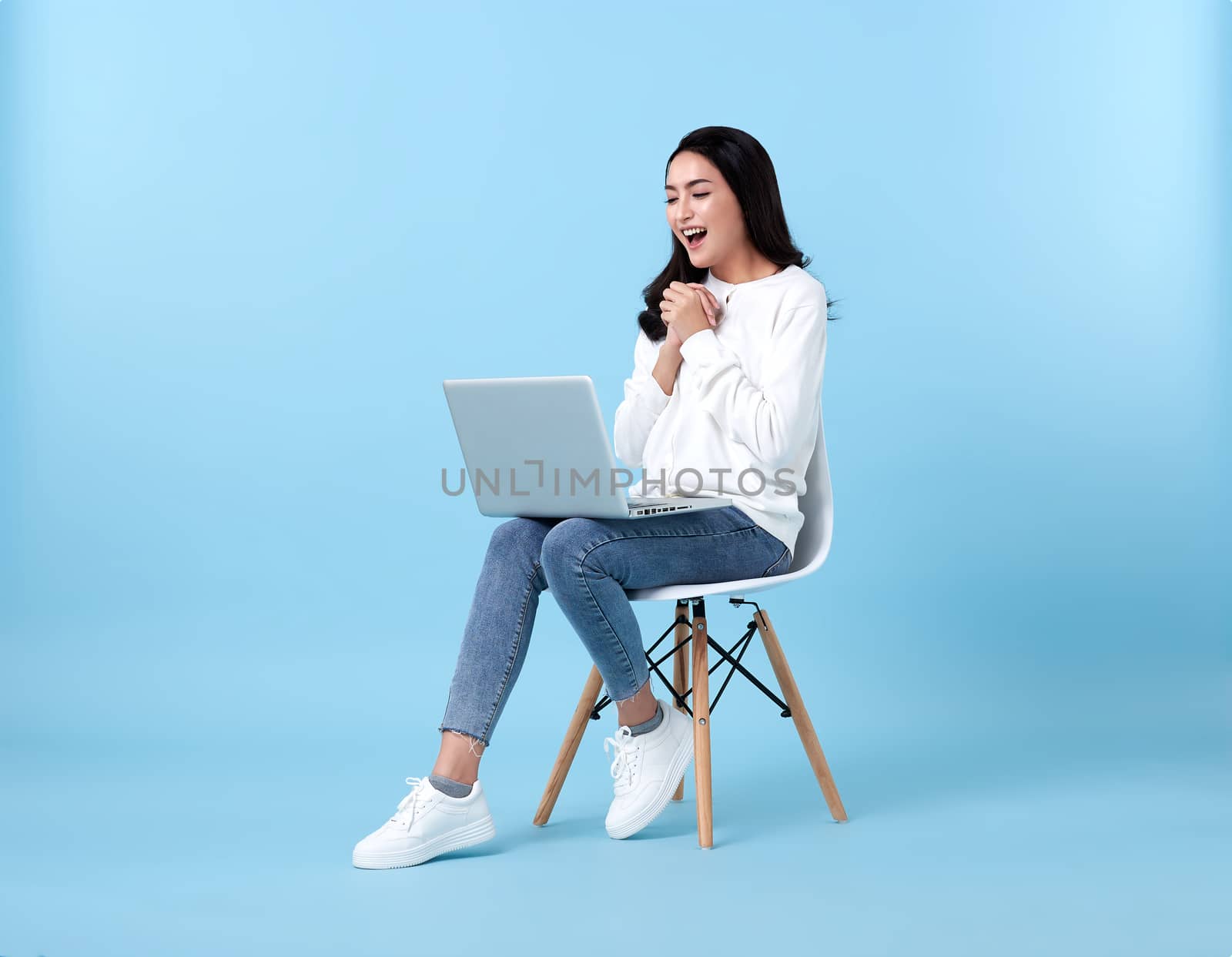 Young woman asian happy smiling in casual white cardigan with denim jeans.While her using laptop sitting on white chair isolate on bright blue background.
